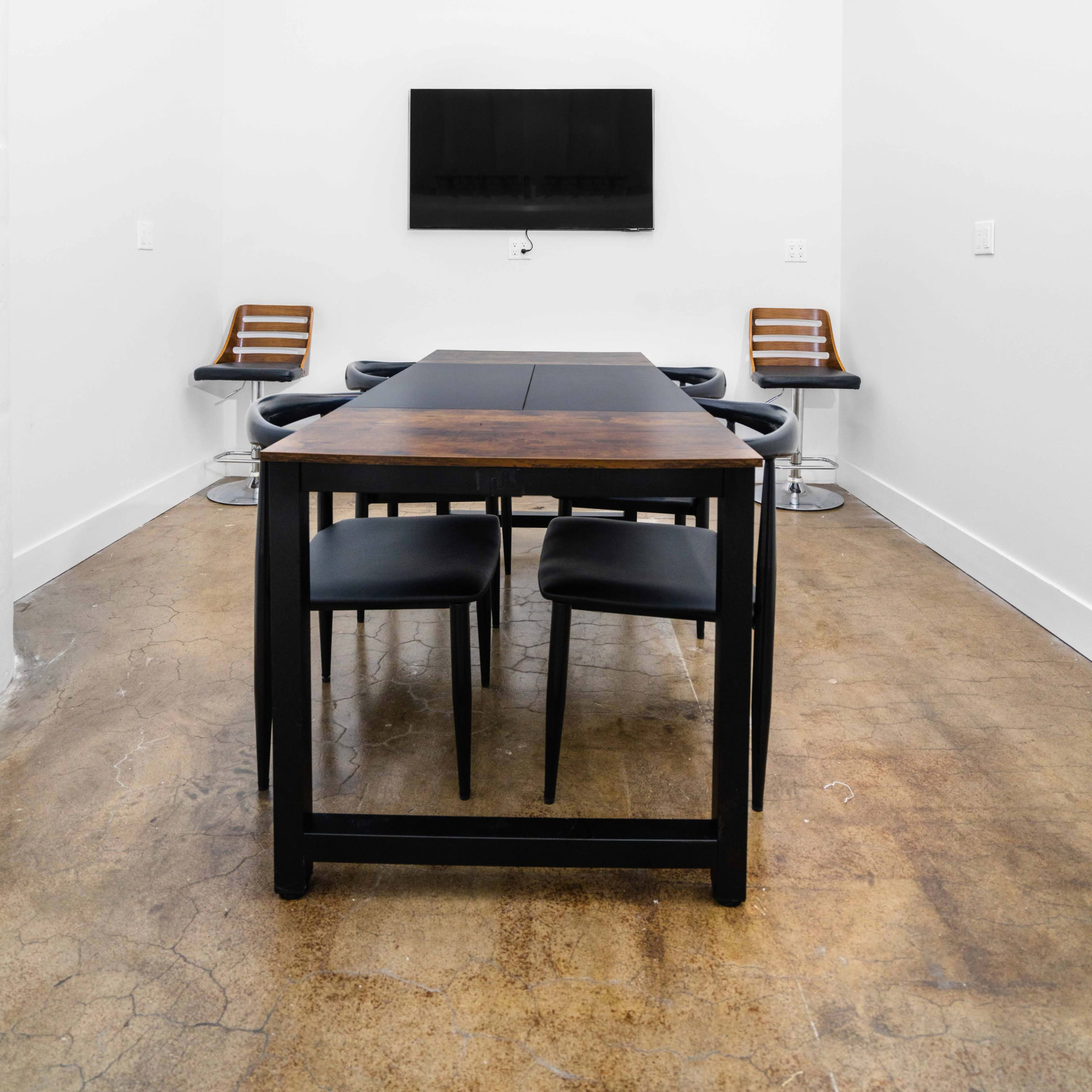 A minimalist meeting room with a black table and chairs, a wall-mounted TV, and concrete flooring.
