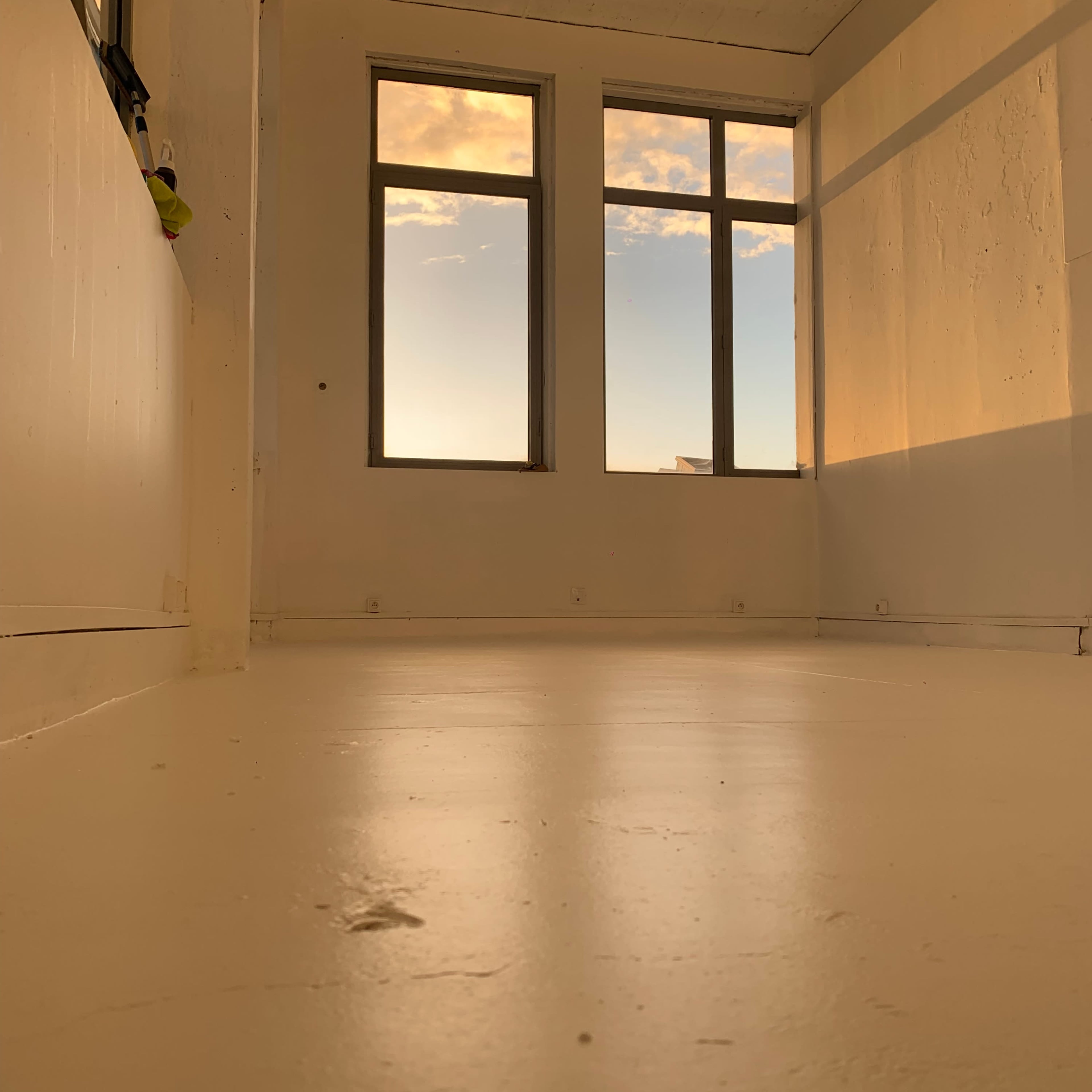 A sparsely furnished room with white walls and large windows reflecting a sunset sky.