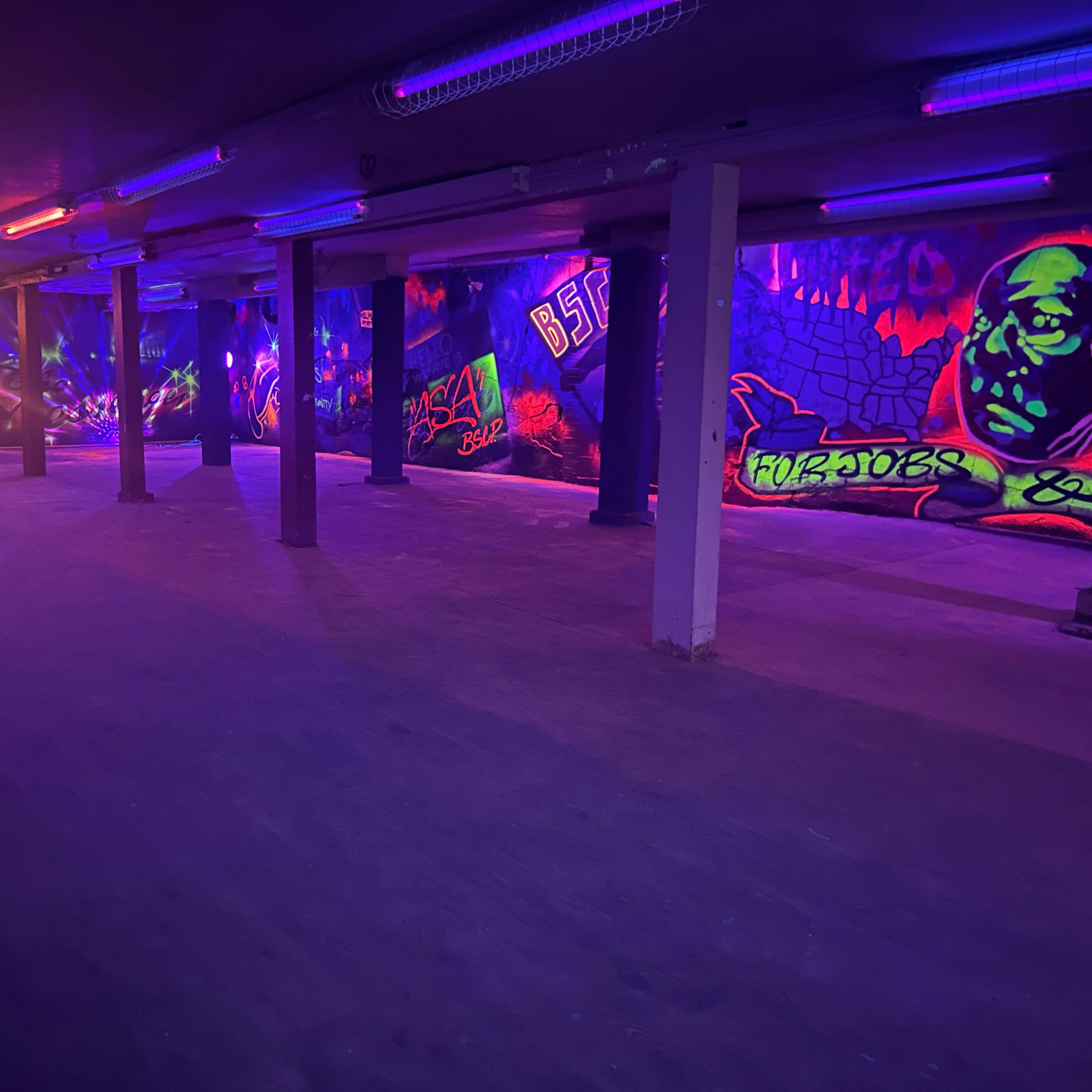 An underground room with vibrant graffiti murals on the walls, illuminated by purple and orange lights.