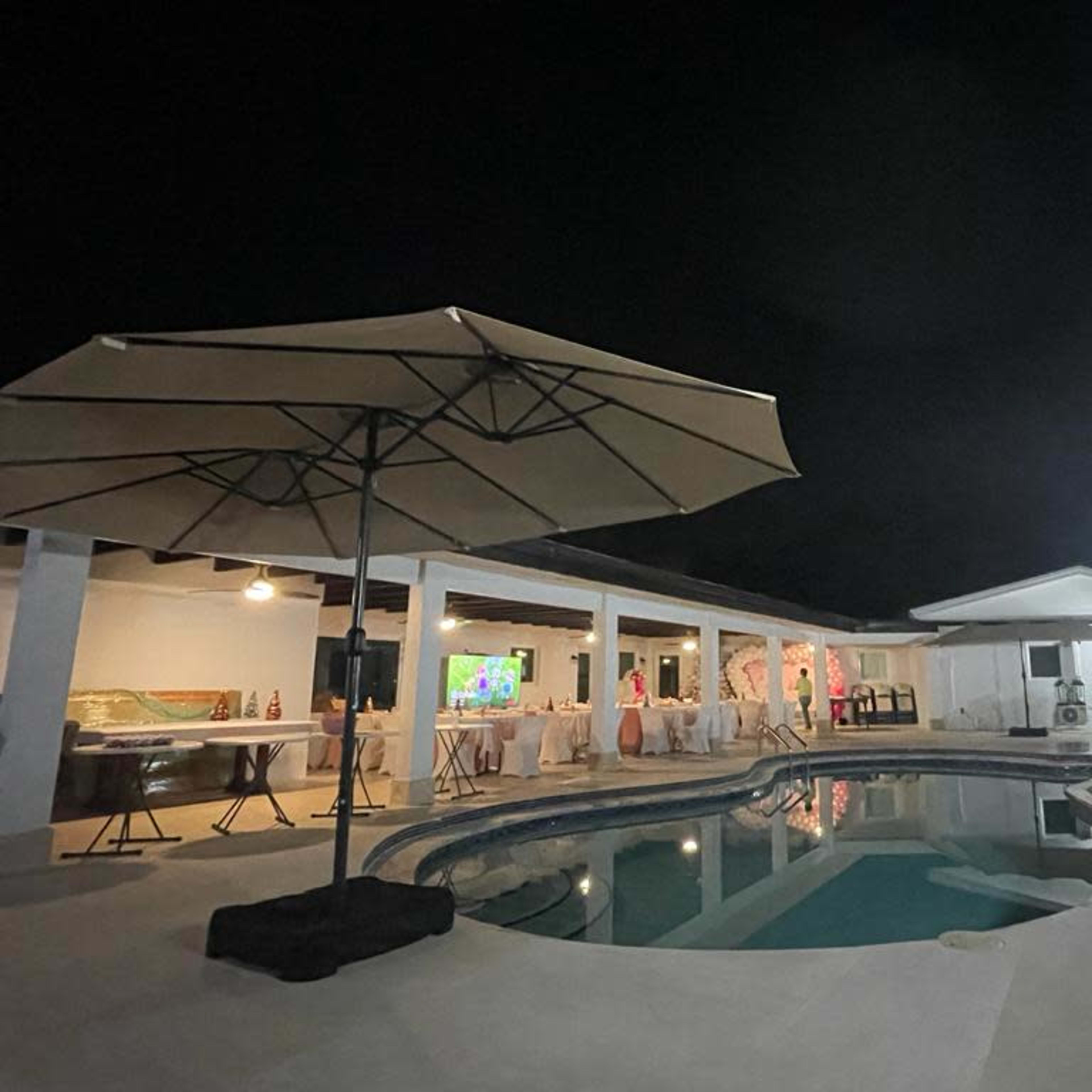 A well-lit backyard features a swimming pool surrounded by a patio, with a large umbrella and a house in the background at night.