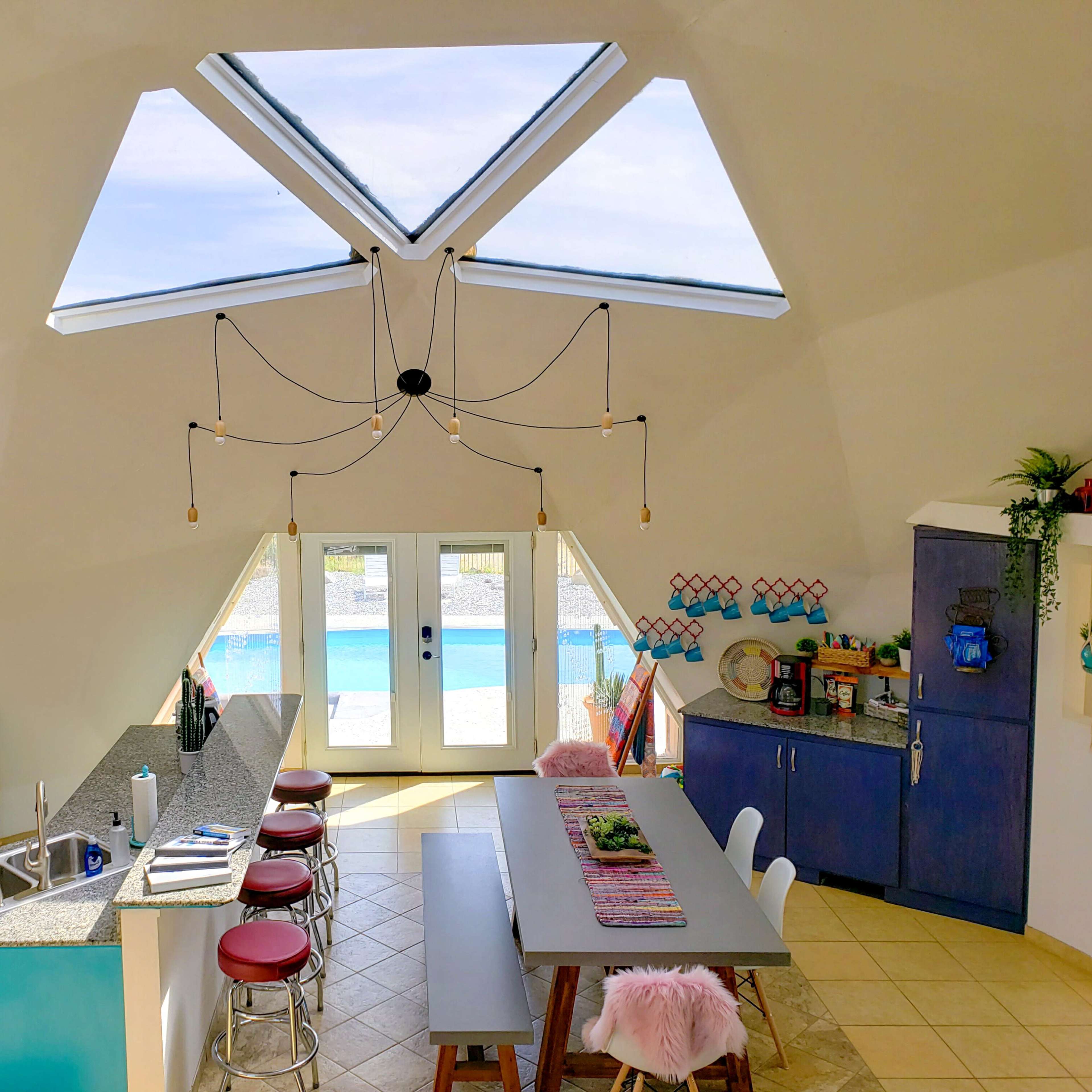 A modern kitchen with a central island, featuring a dining table, large windows, and a pool visible outside.