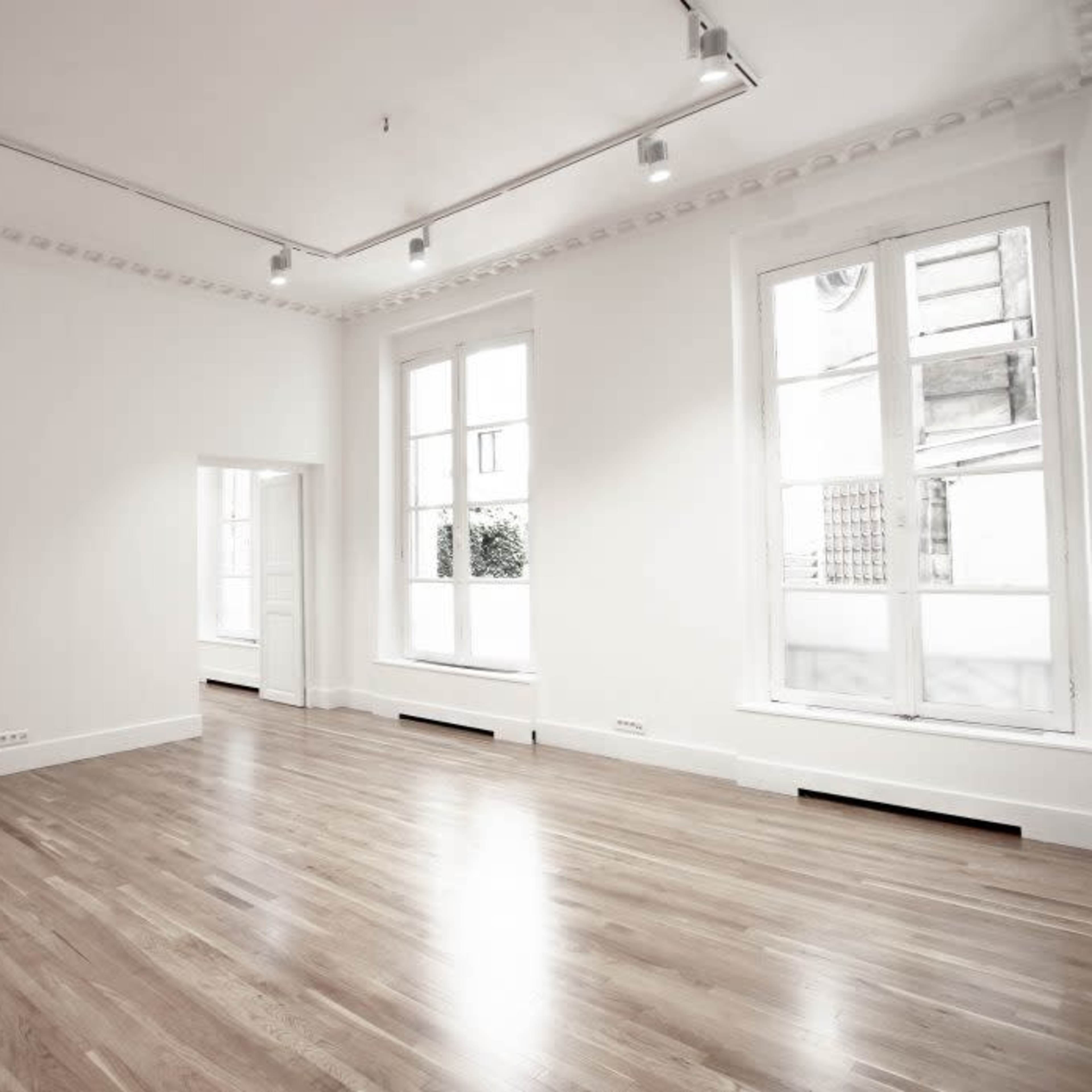 A spacious, empty room with wooden flooring, large windows, and white walls.