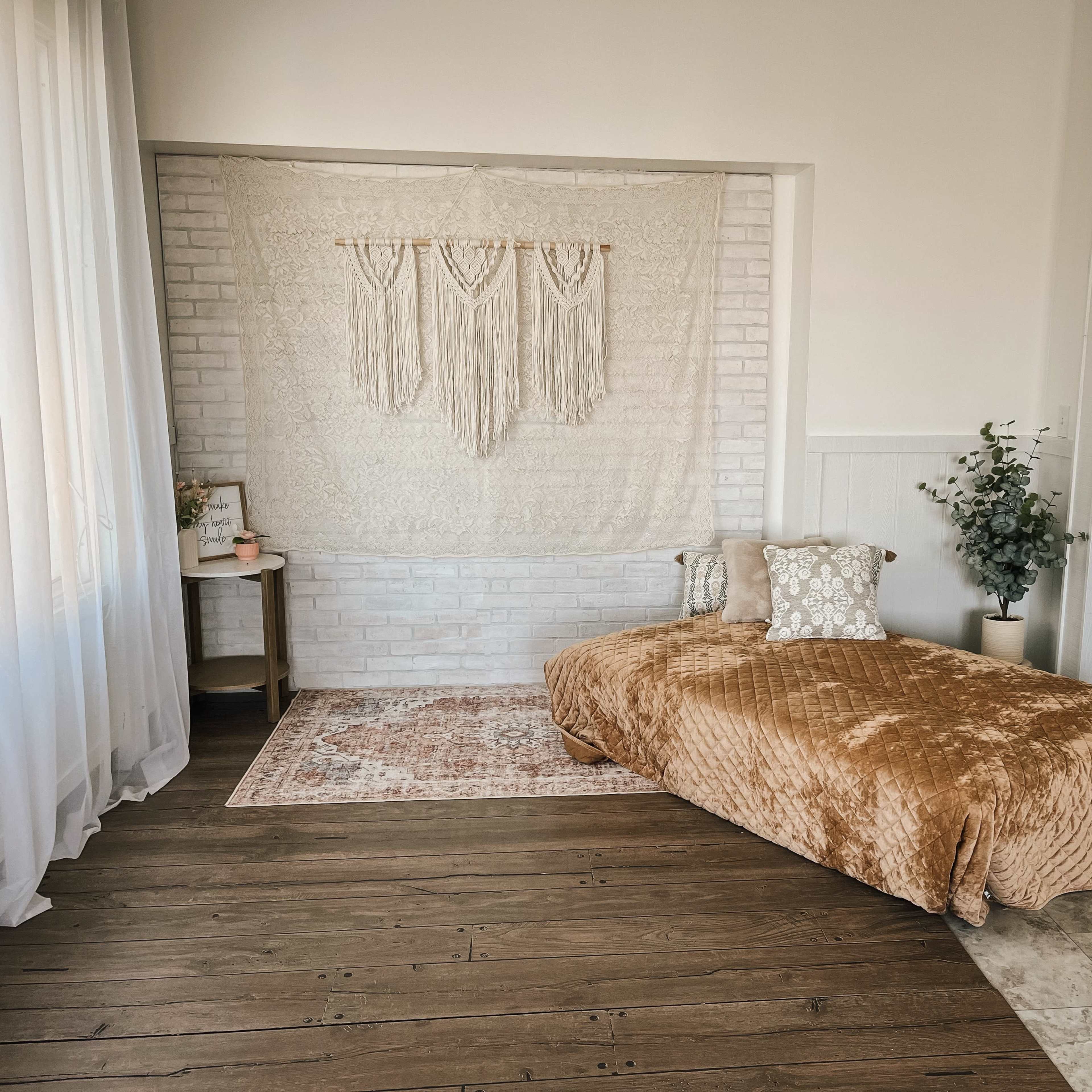 A bright, minimalist bedroom with a macramé wall hanging, a daybed covered in a textured blanket, and a small side table next to a potted plant.