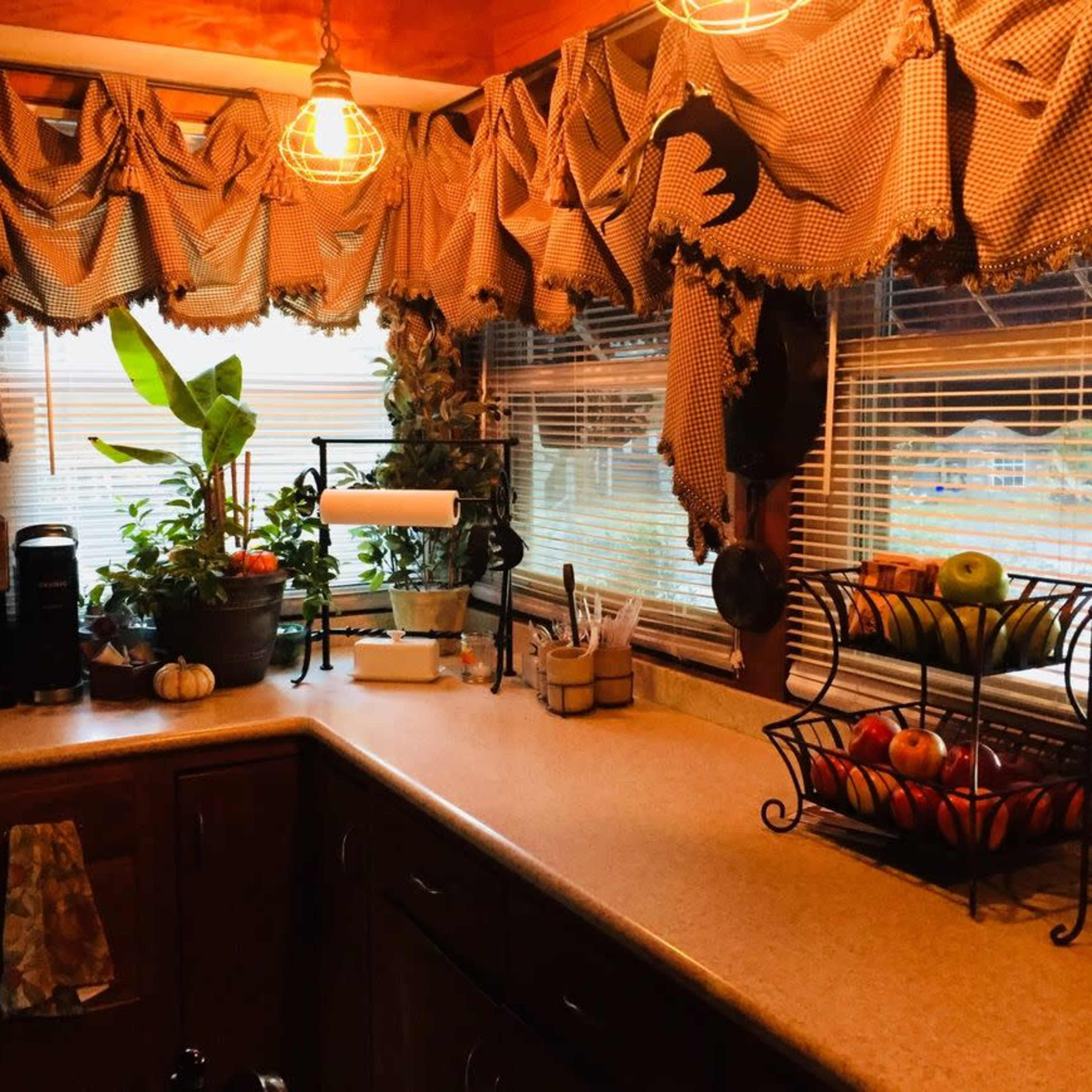 A cozy kitchen with wooden cabinets, a countertop adorned with a fruit basket, and a window dressed in checkered curtains, allowing natural light to enter.