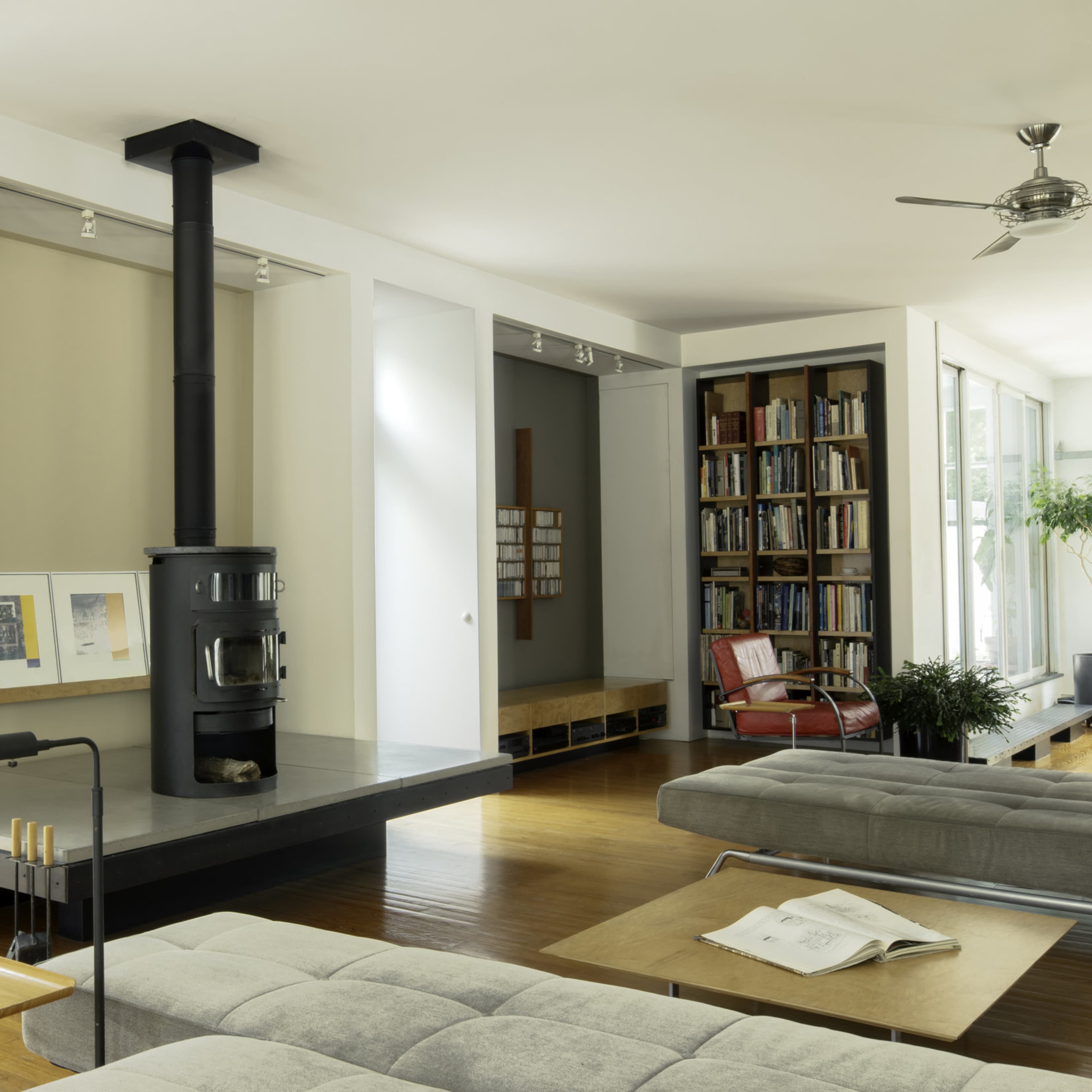 A modern living room features a prominent black wood stove, large windows, and sleek furniture arranged around a hardwood floor.