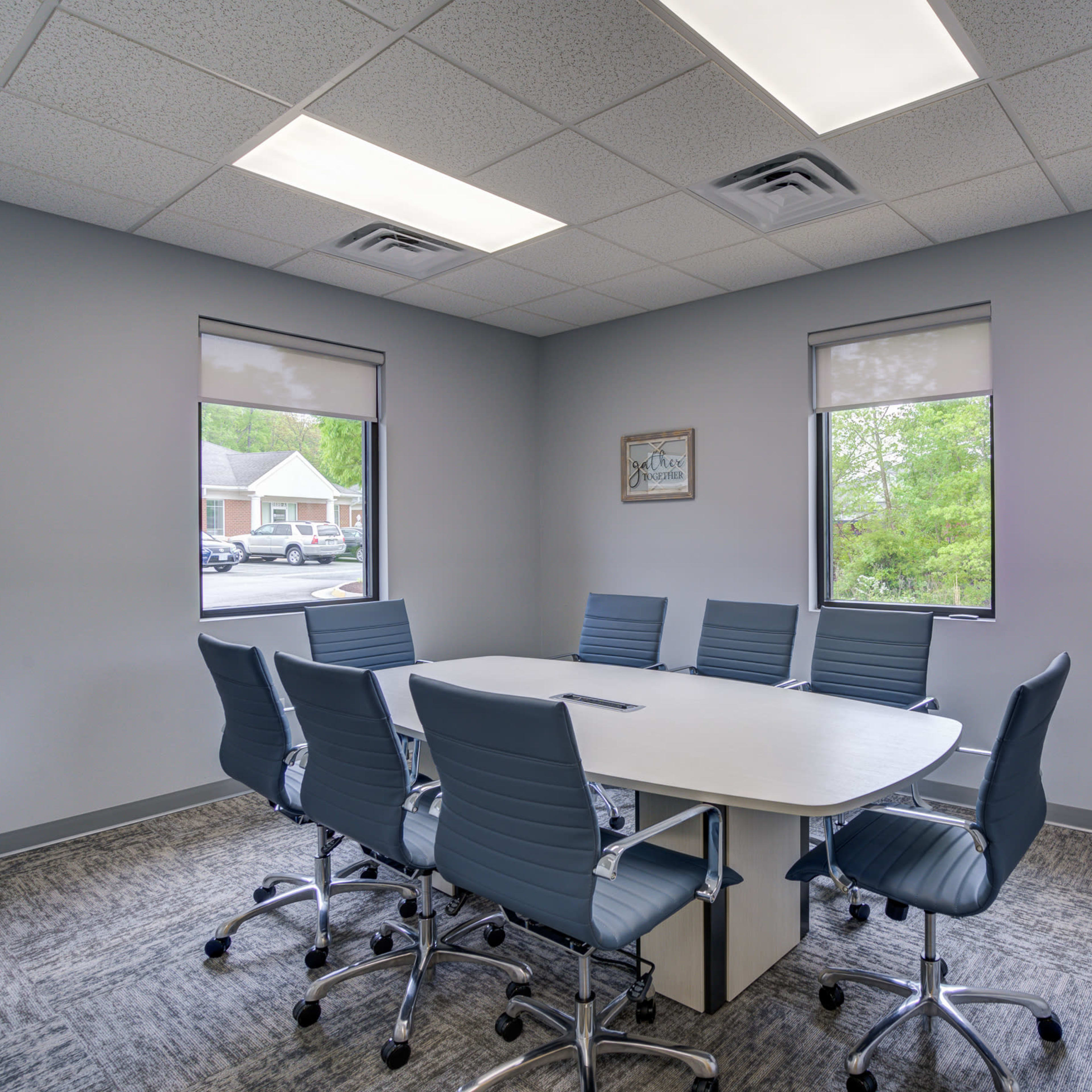 A conference room with a large white table surrounded by eight blue chairs and two windows letting in natural light.