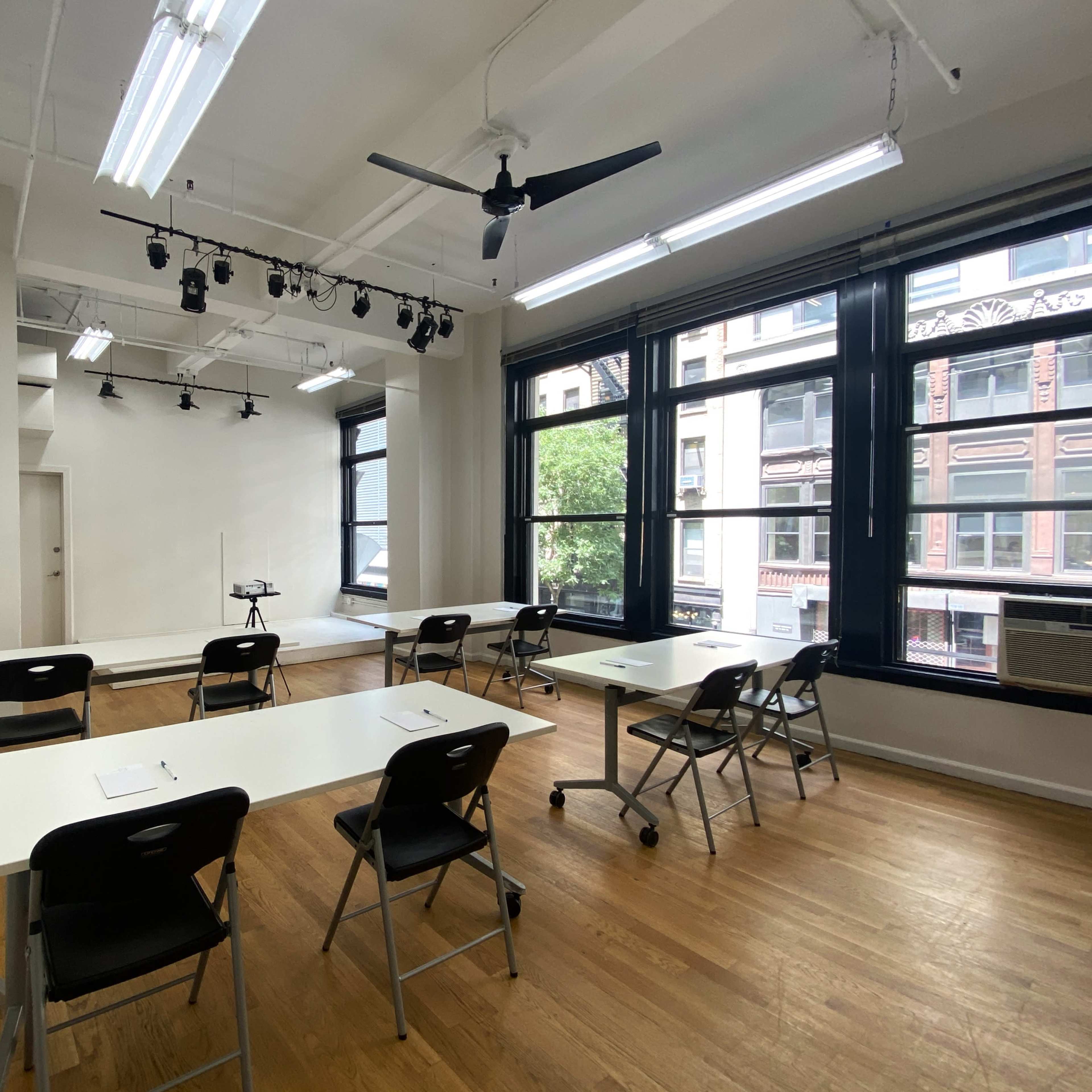 A bright classroom with white tables, black chairs, and large windows overlooking a city street.