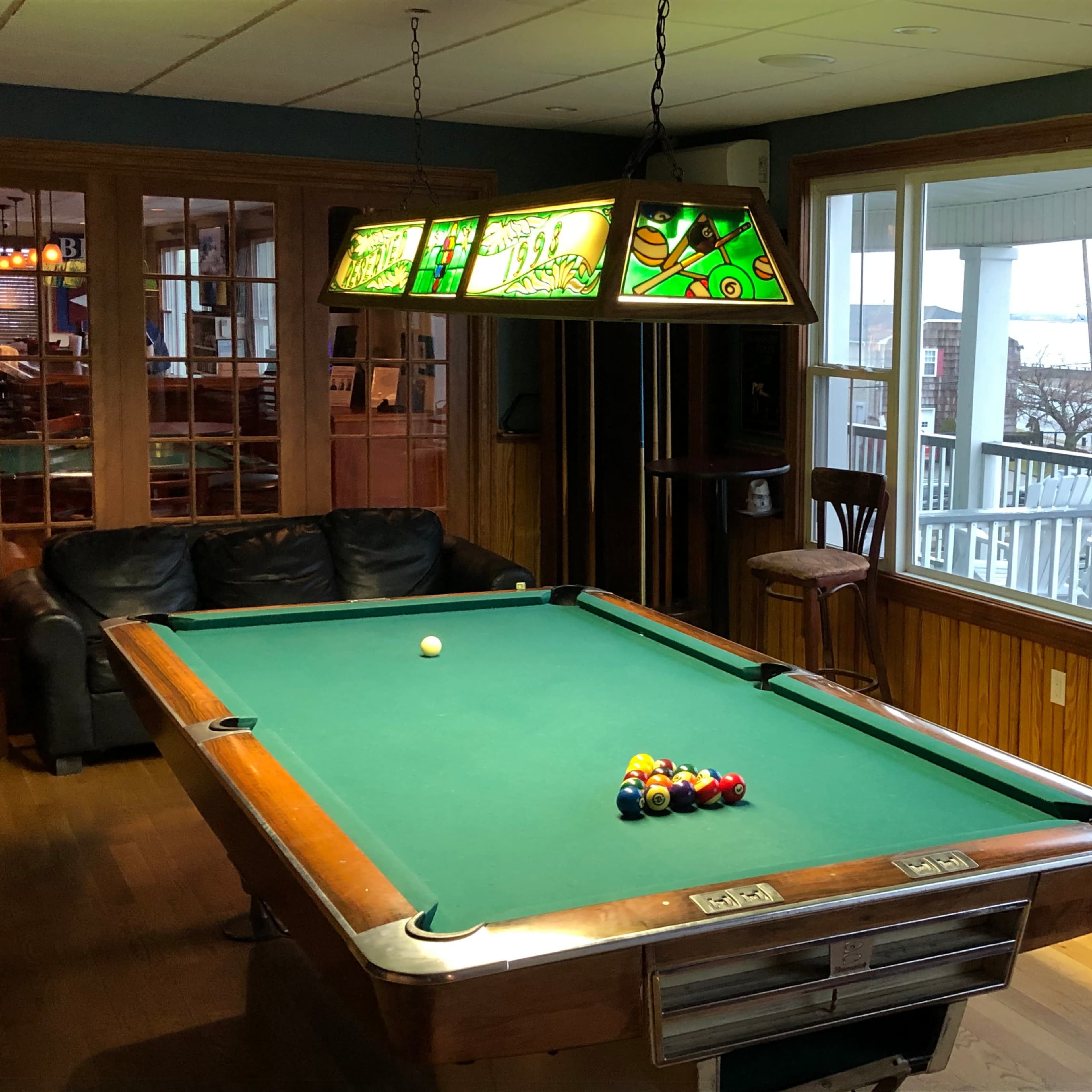 A pool table with a set of billiard balls arranged on it is located in a room with large windows overlooking a body of water.