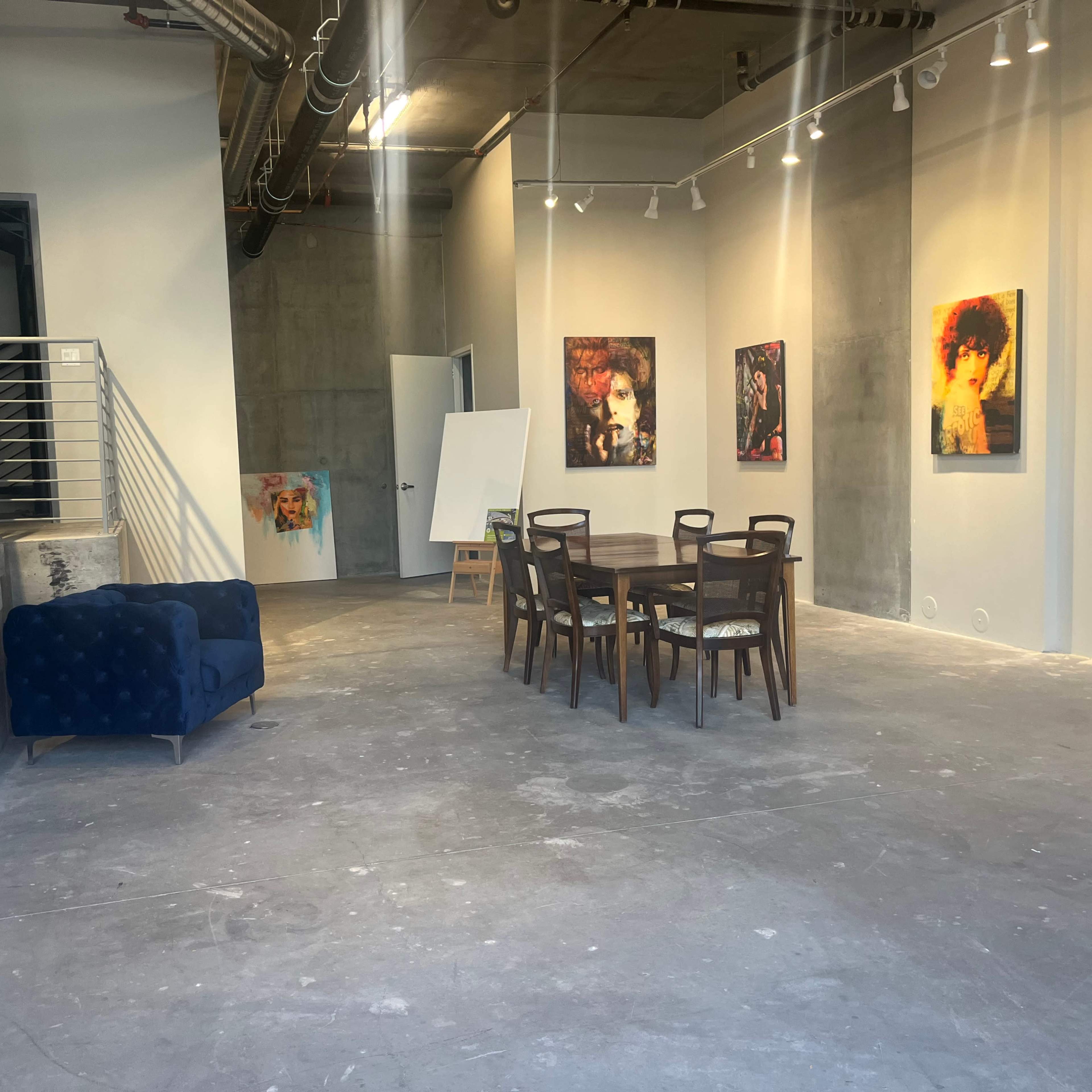 A minimalist art gallery with a staircase on the left, a blue couch, a wooden dining table surrounded by chairs, and various artworks displayed on the walls.