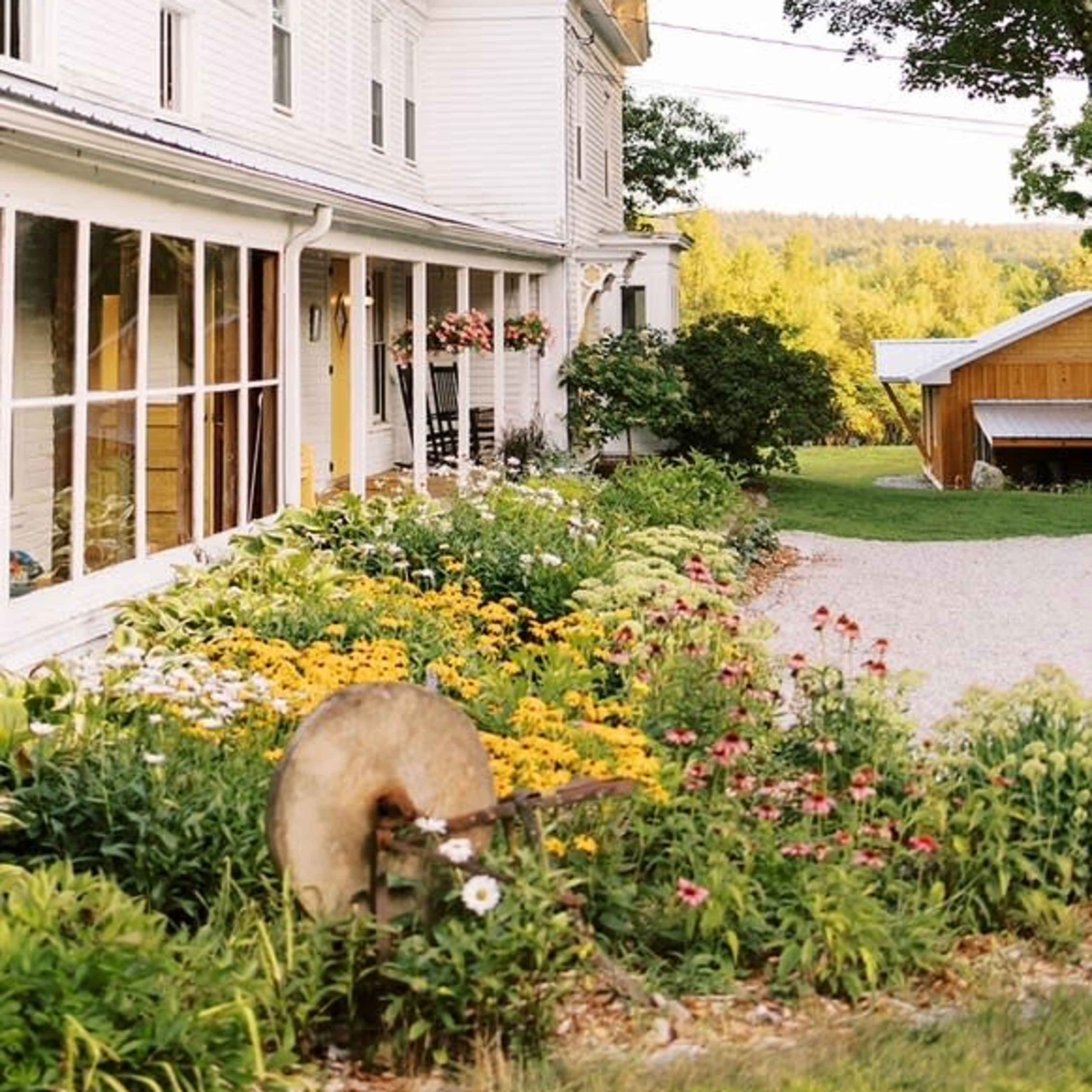 A white farmhouse with a metal roof is set behind a colorful flower garden and gravel driveway, surrounded by trees in a rural landscape.