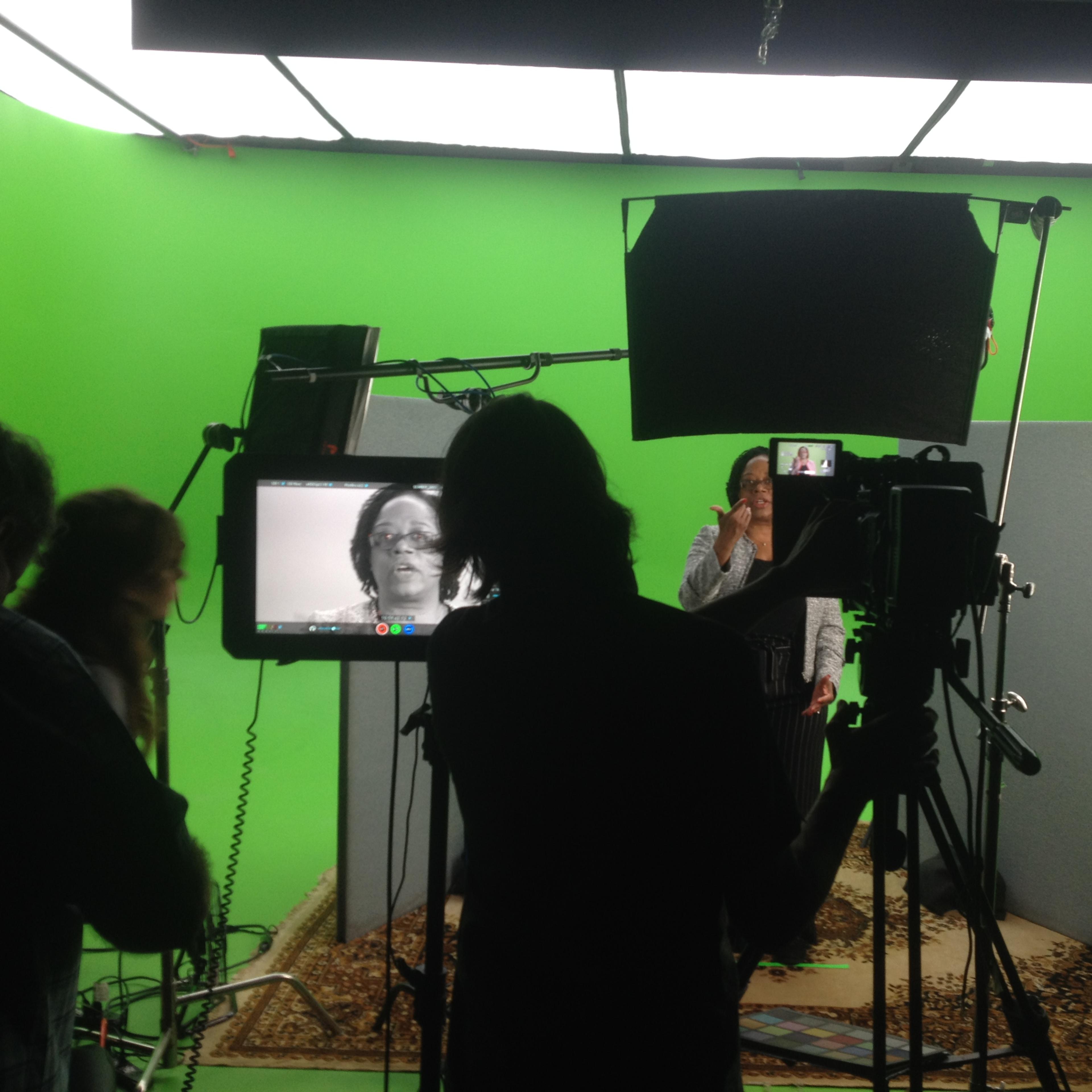 A person stands in front of a green screen while being filmed by multiple cameras and crew members.