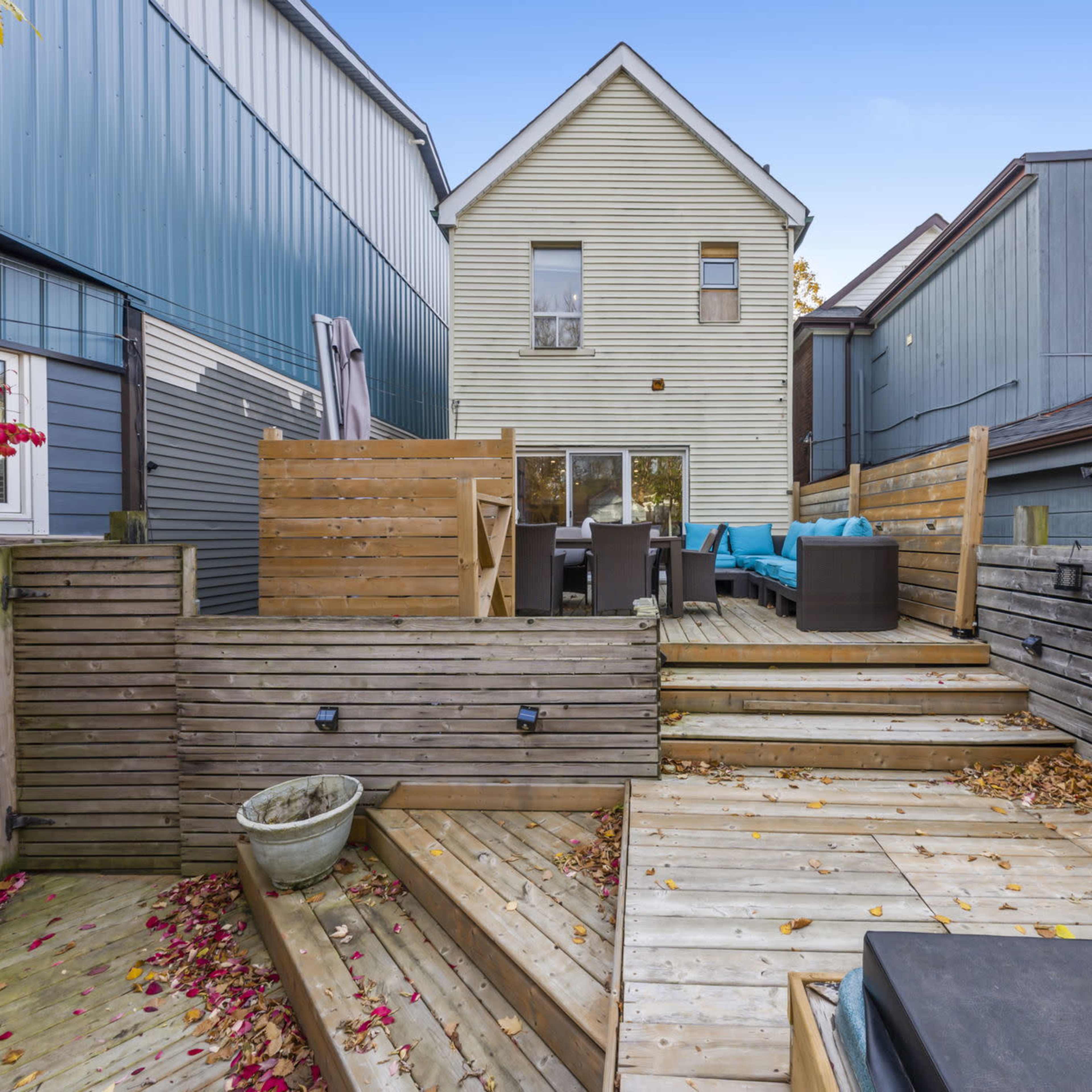 A backyard patio with wooden decking, featuring seating areas, a plant pot, and buildings in the background.