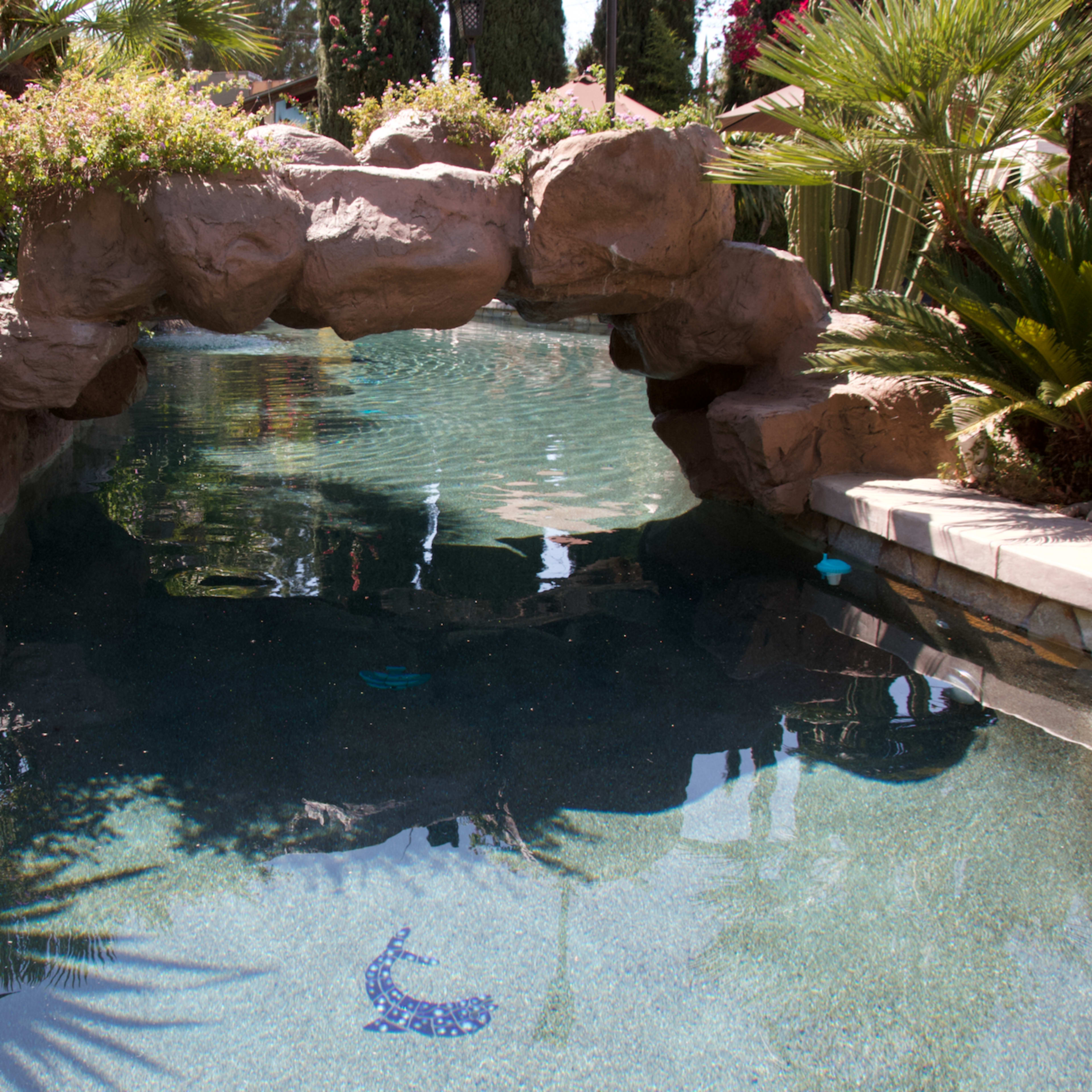 A swimming pool with a stone arch bridge surrounded by lush greenery and colorful flowers.