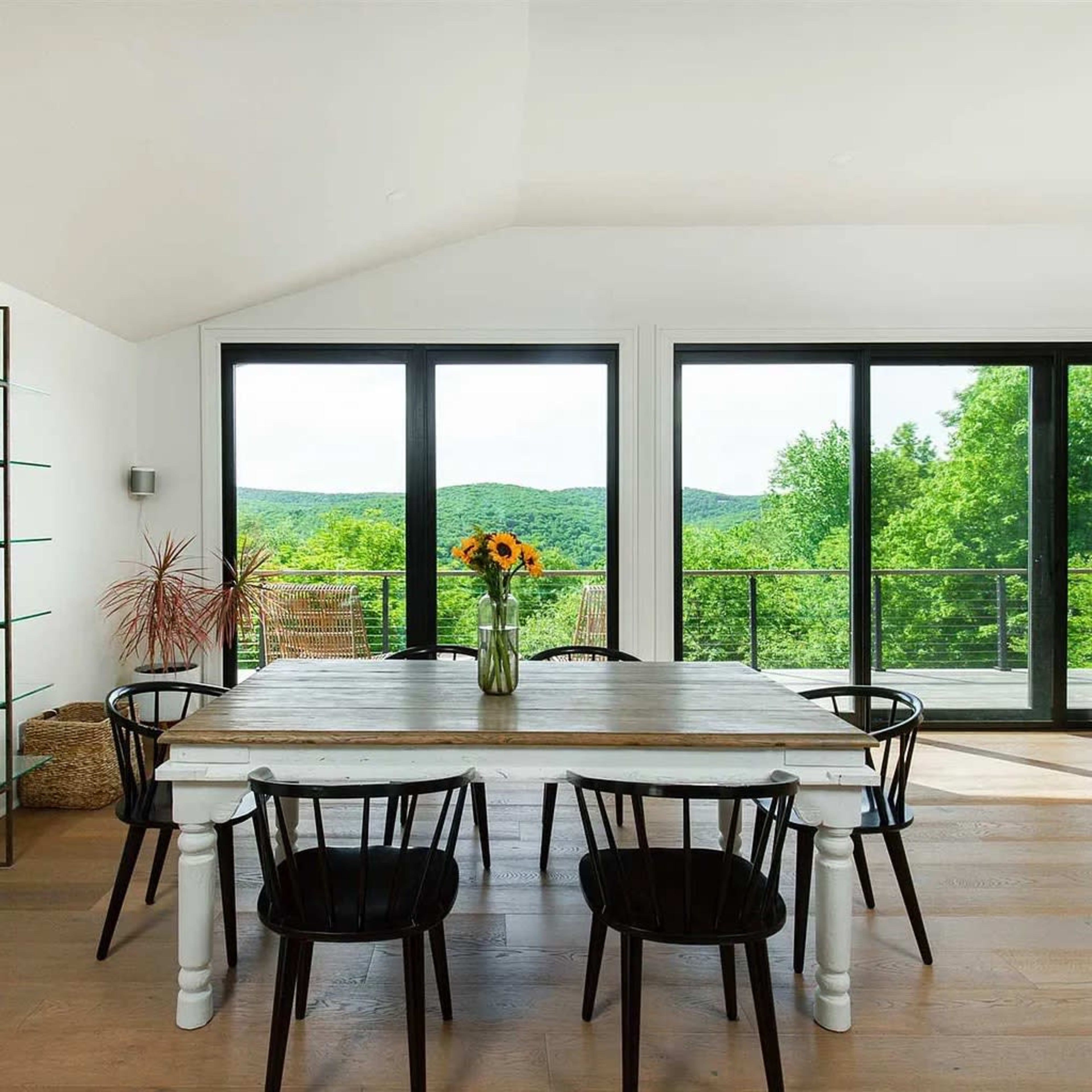 A dining area features a wooden table with black chairs, surrounded by large windows that showcase a view of green hills.