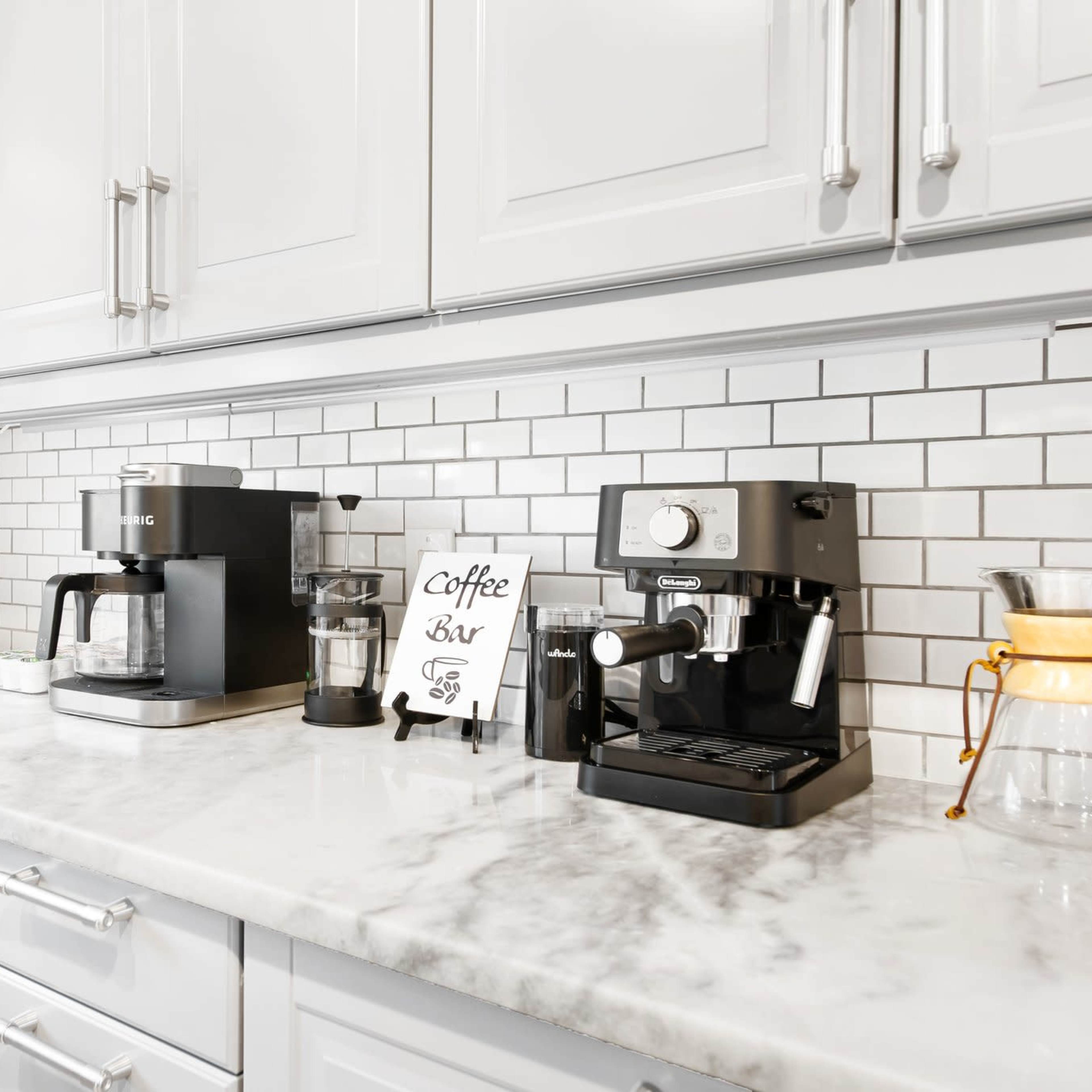 A kitchen countertop features a coffee bar with various coffee machines, a toaster, and a decorative sign amidst a white and gray tiled backdrop.
