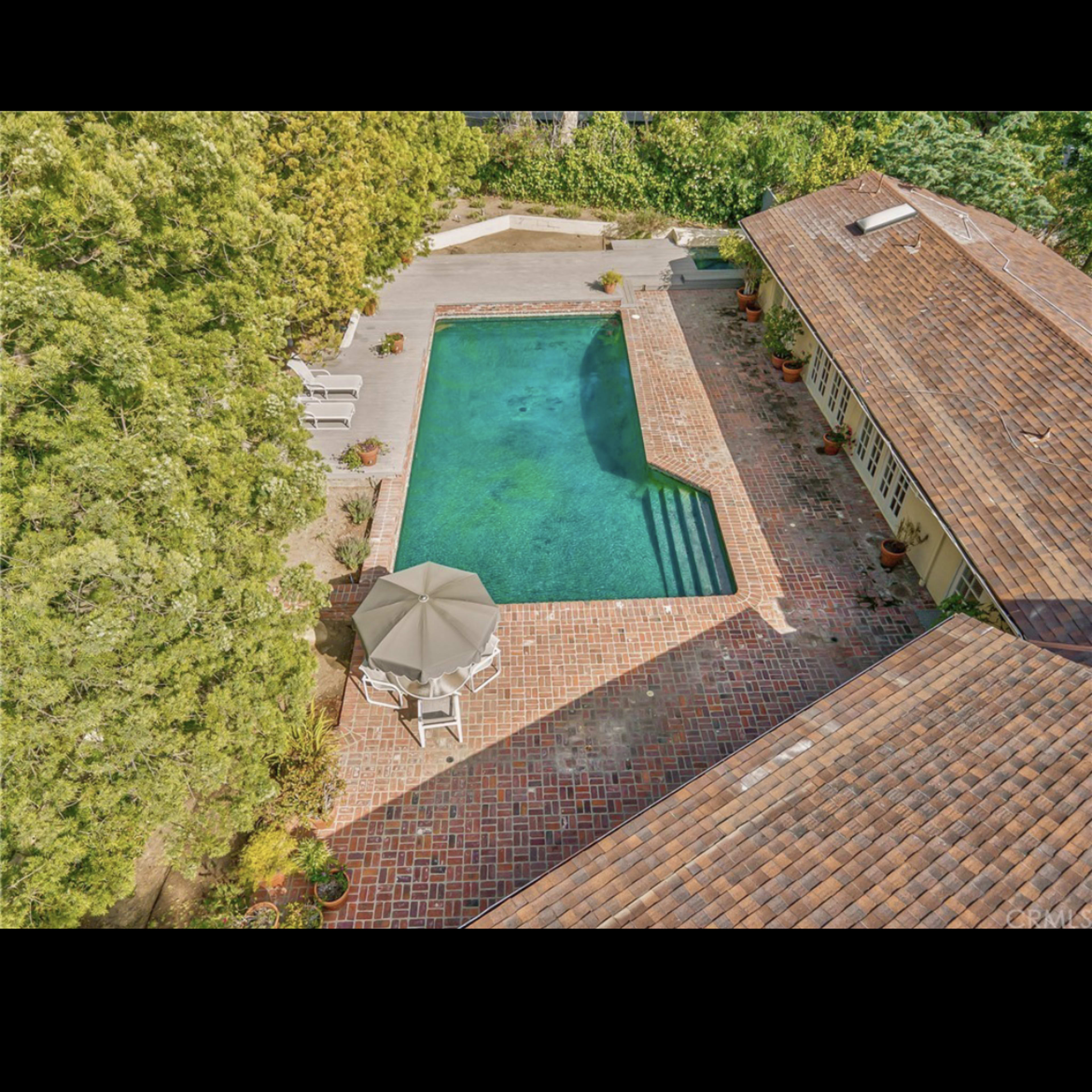 An aerial view shows a rectangular swimming pool surrounded by a brick patio and a shaded umbrella area, adjacent to a house with a tiled roof.