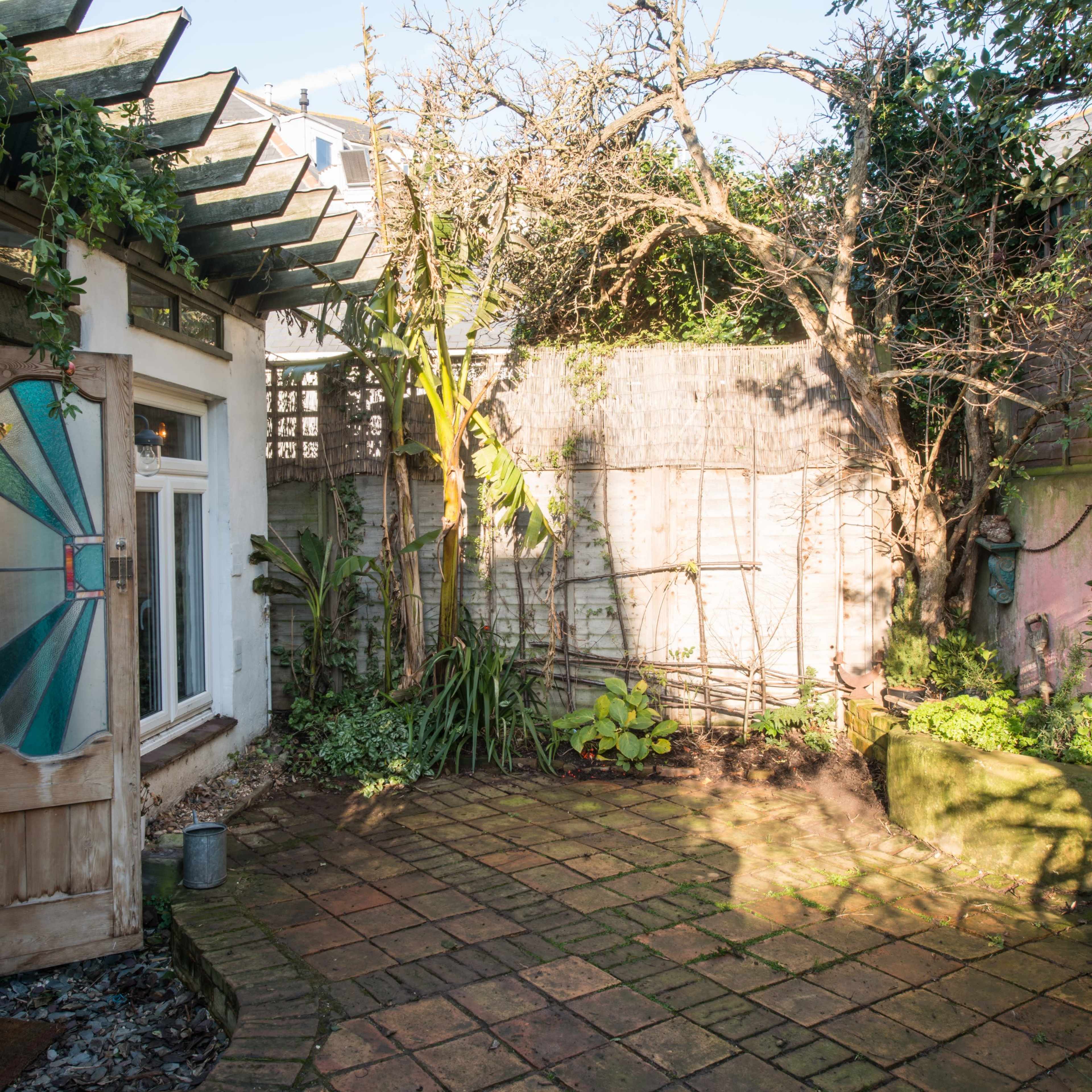 A small, sunlit garden with a stone patio, surrounded by plants and a wall covered in greenery.