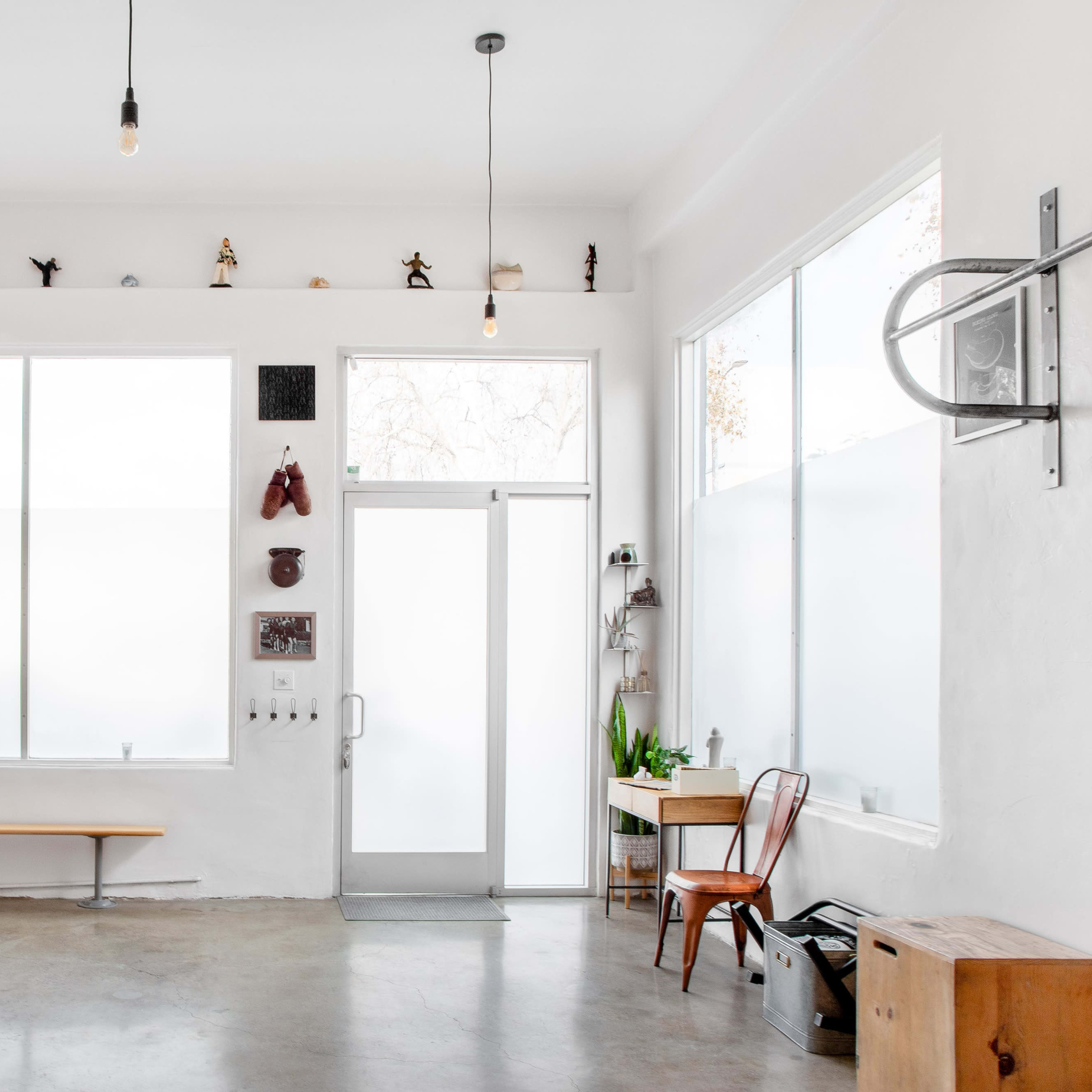 A bright, minimalistic interior studio with a wooden bench, large windows, and various decorative items on the walls.