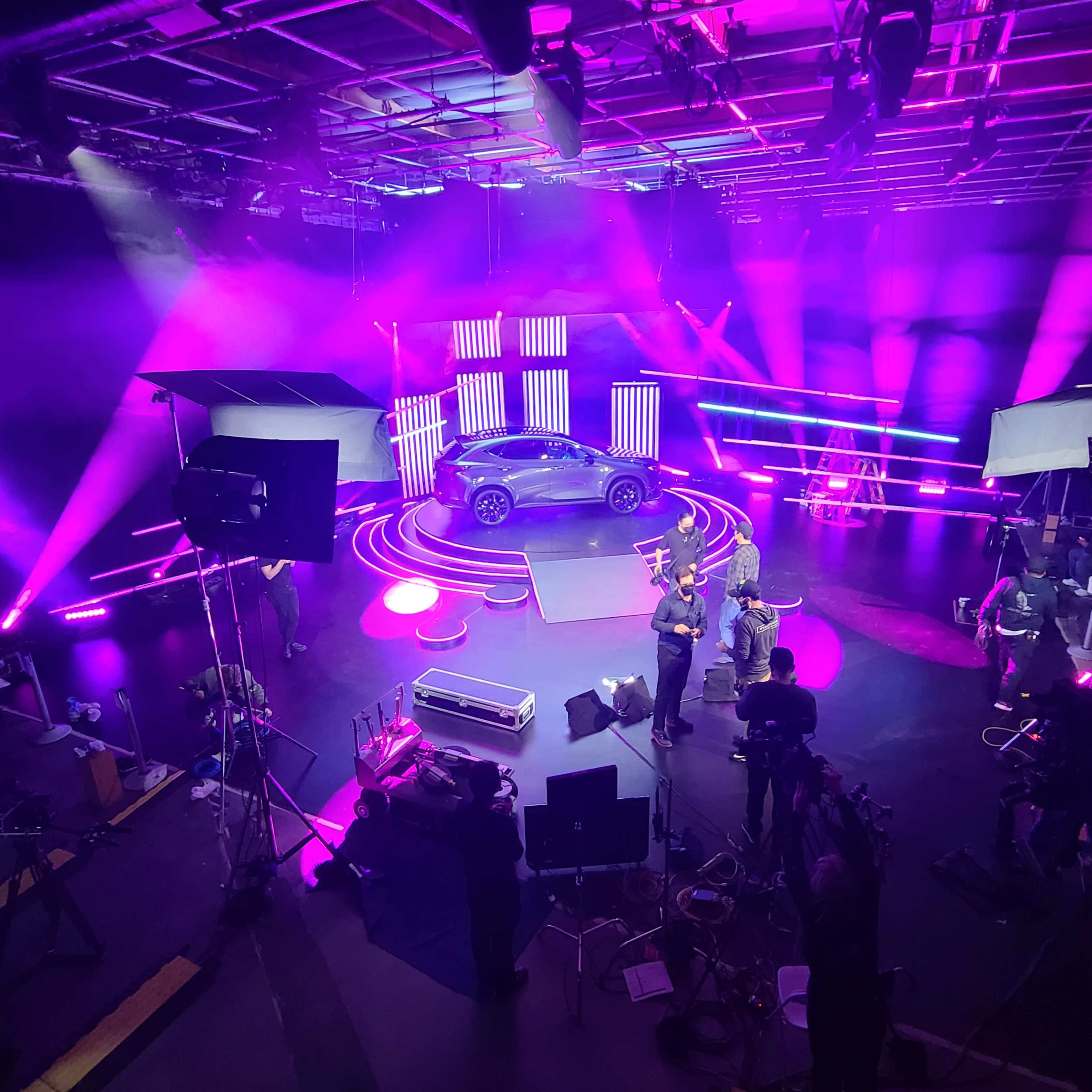 A sleek car is showcased on a circular platform surrounded by vibrant lights and film equipment in a studio setting.