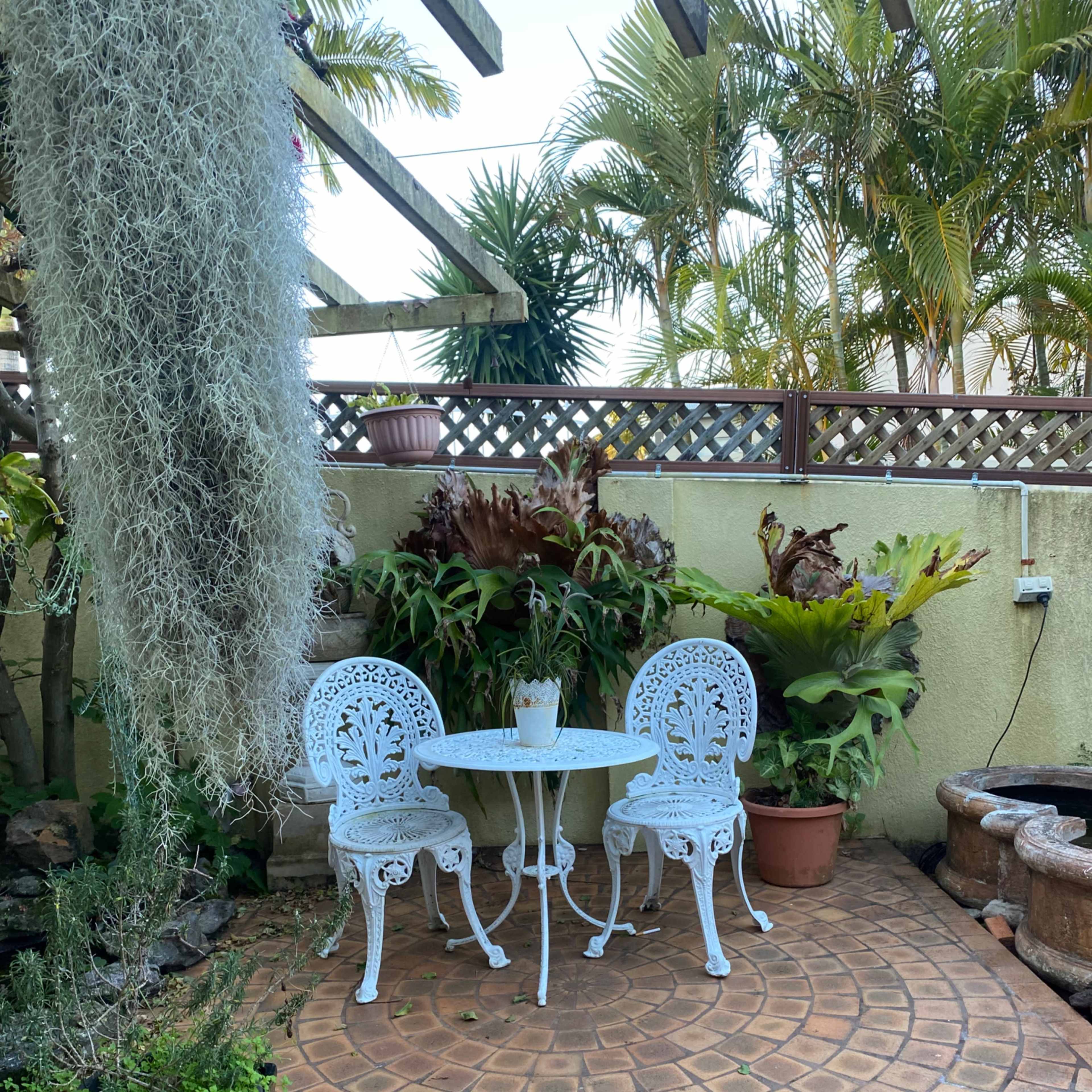 A small outdoor patio features a round table and two ornate white chairs surrounded by greenery and planters.