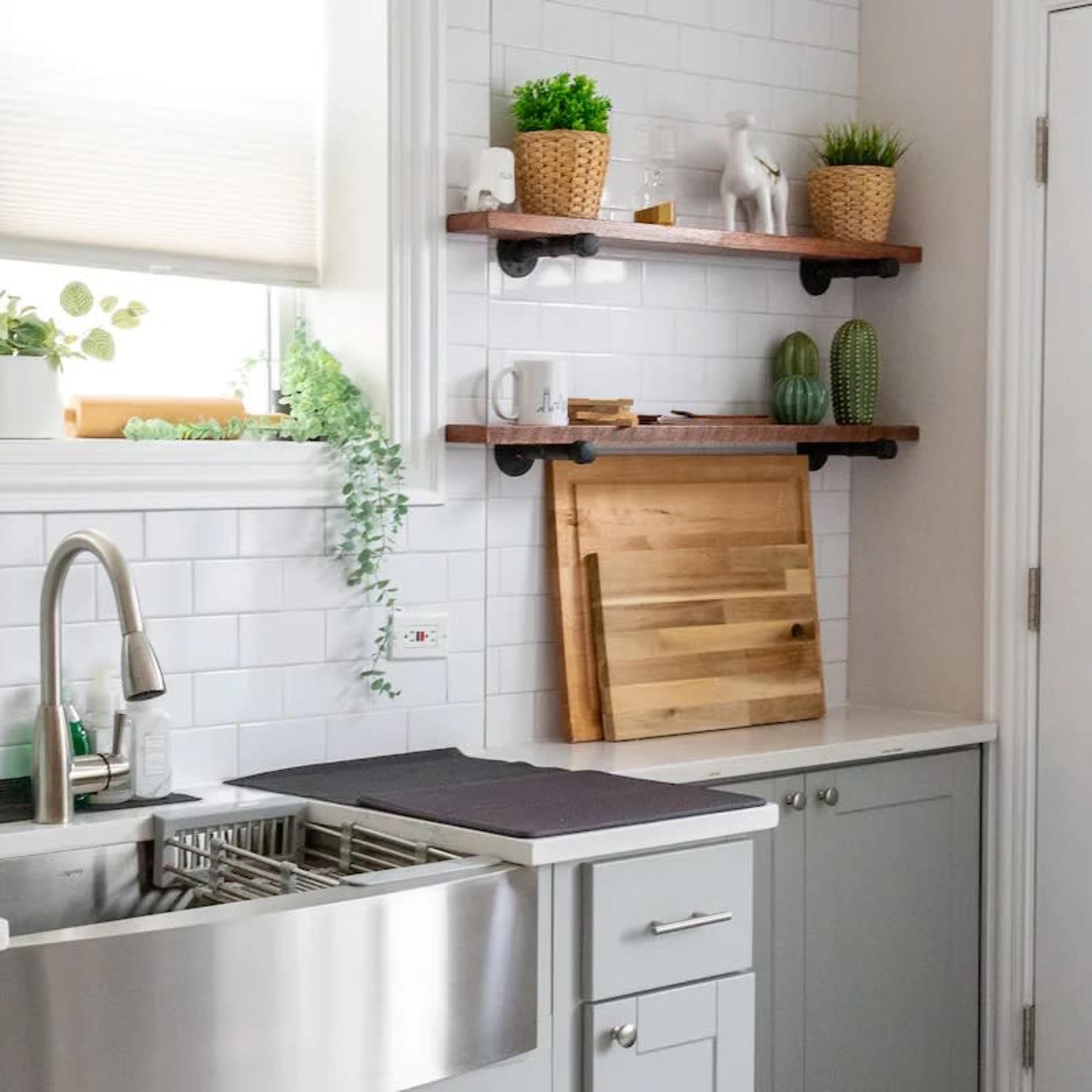 A modern kitchen features a stainless steel sink, white tiled walls, and wooden shelves holding decorative plants and kitchen items.