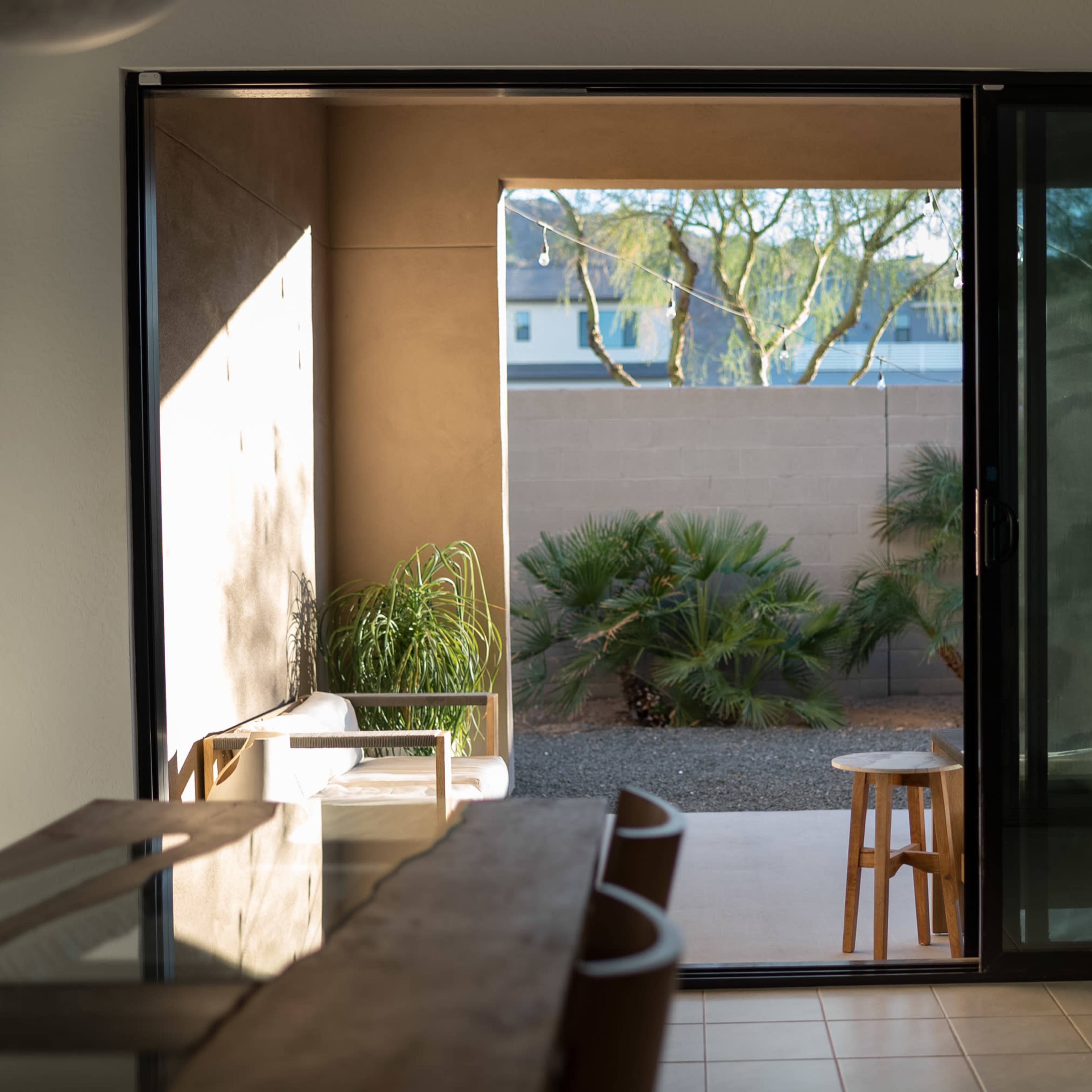 A sliding glass door opens to a patio with a small stool and potted plants, framed by a wall and trees beyond.