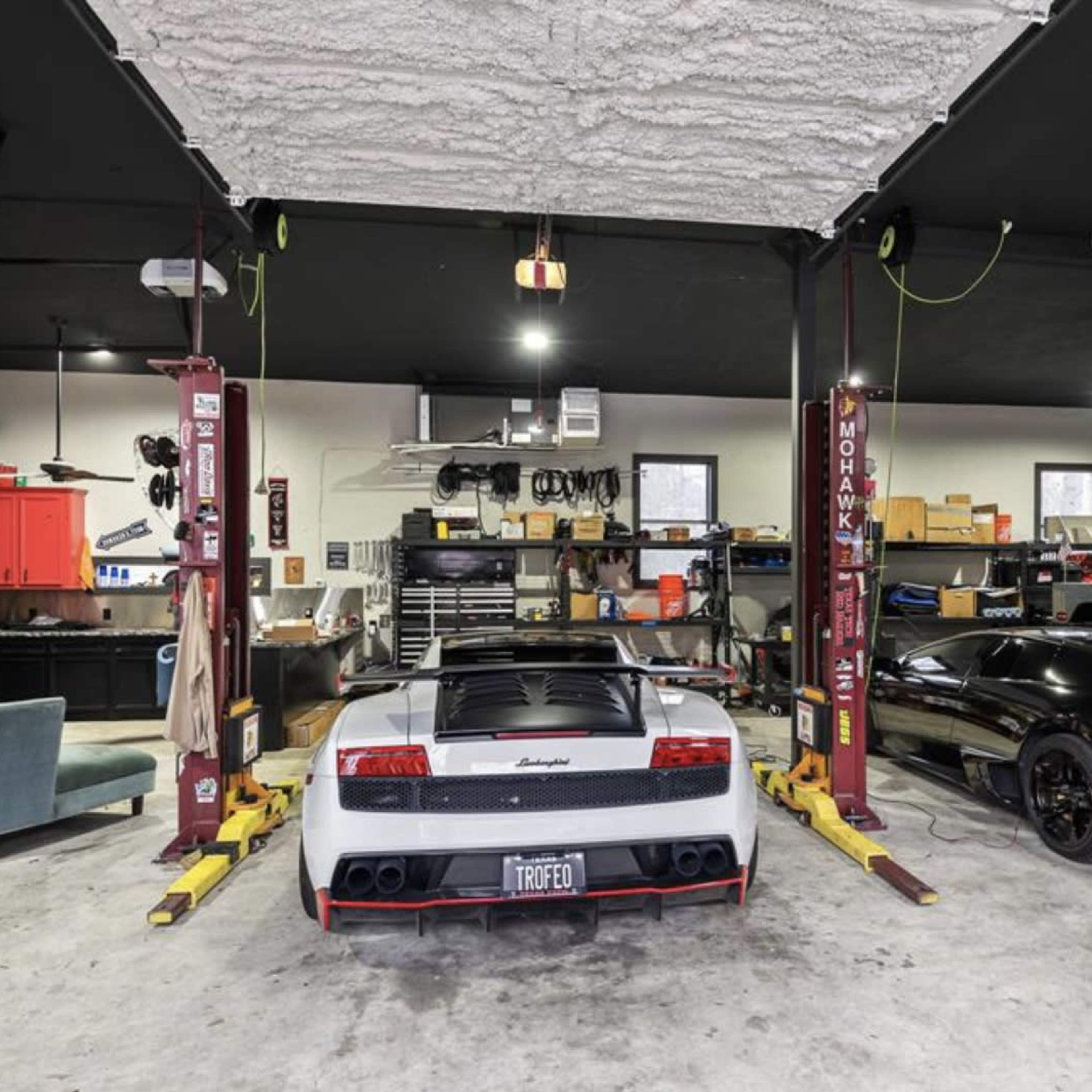A garage with two luxury sports cars, one white and the other black, positioned on lifts amidst a cluttered workspace featuring tools and equipment.