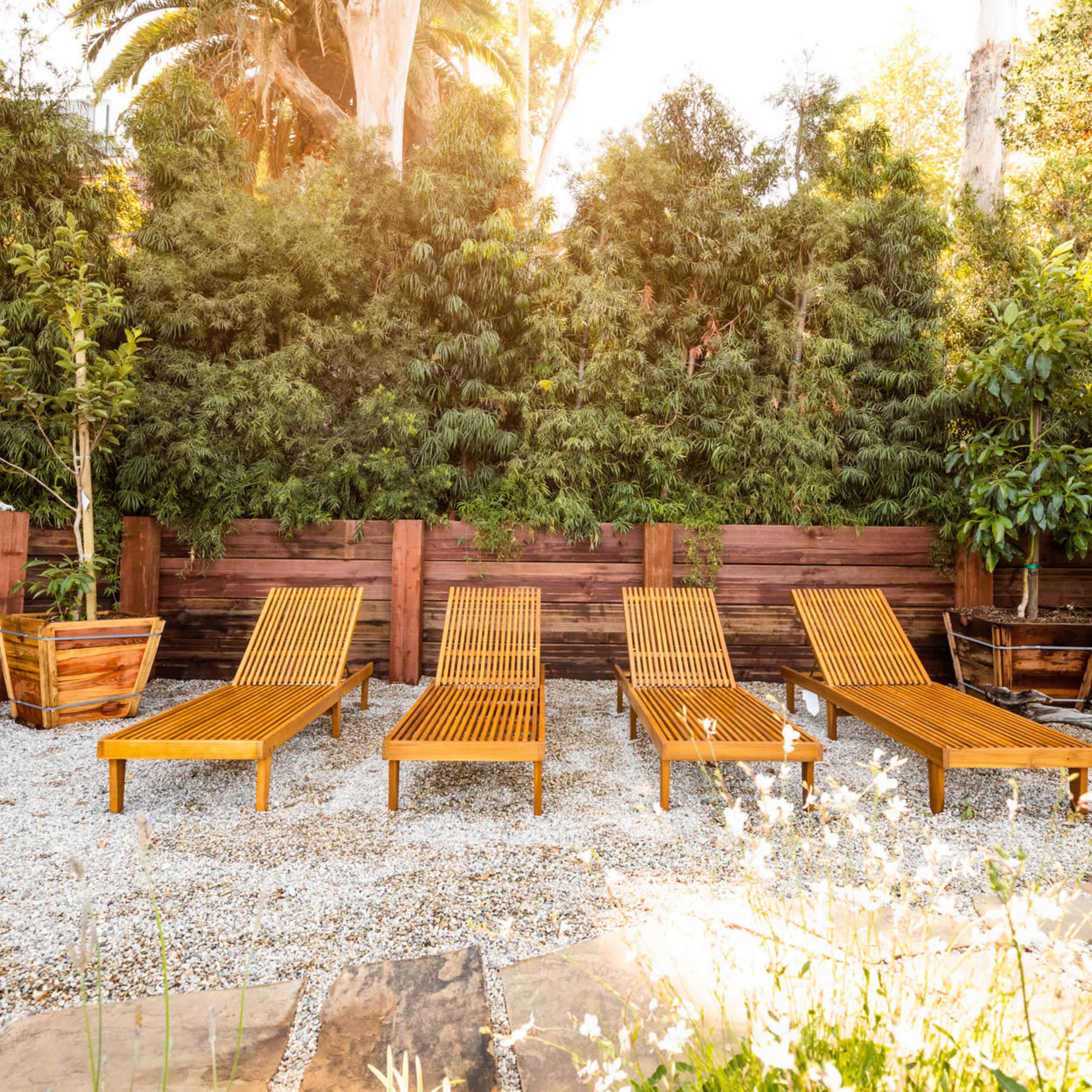 A serene outdoor space featuring four wooden lounge chairs positioned on a gravel area surrounded by lush greenery and wooden planters.