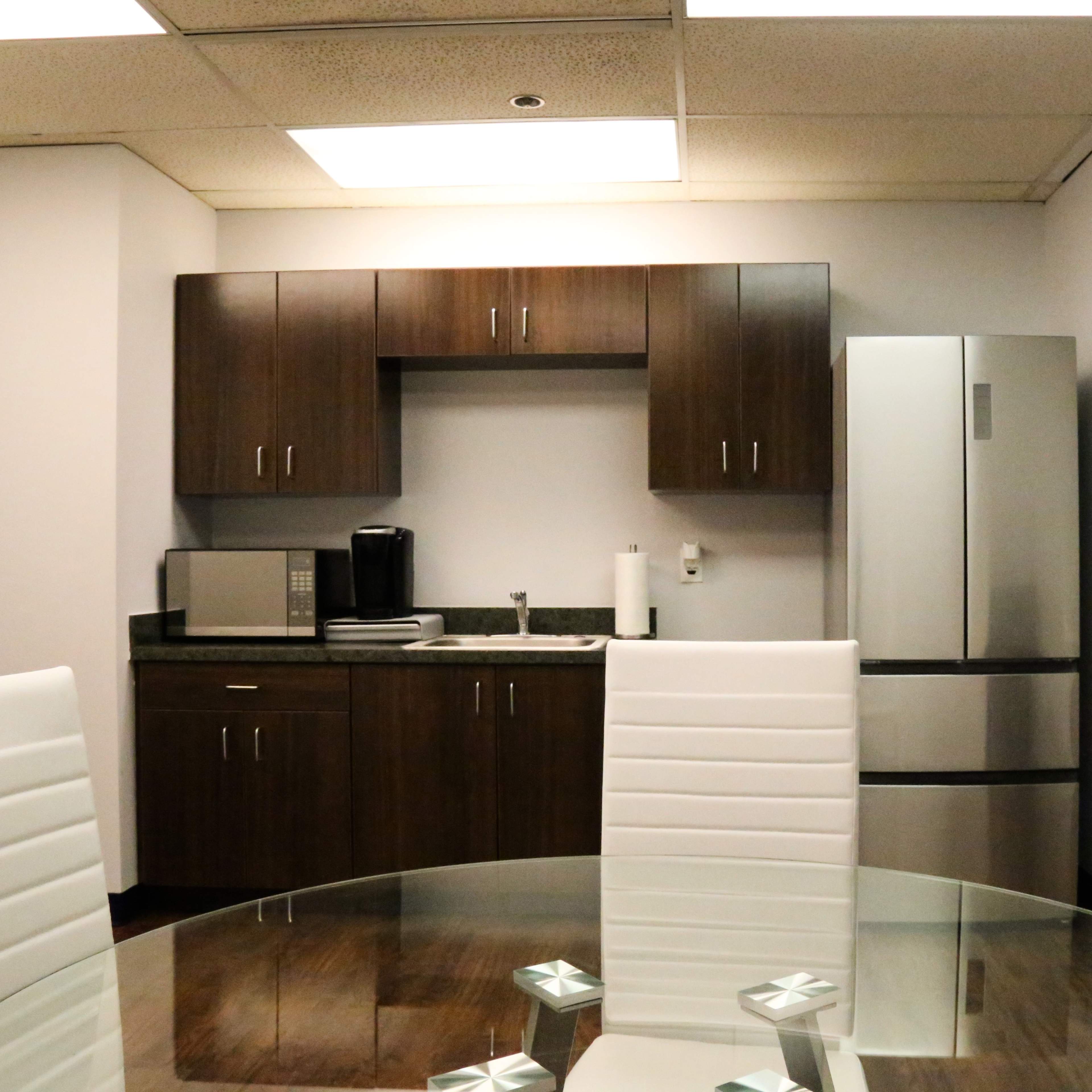 A modern kitchenette with a microwave, coffee maker, and a refrigerator, set against a backdrop of light-colored walls and a glass dining table with white chairs.