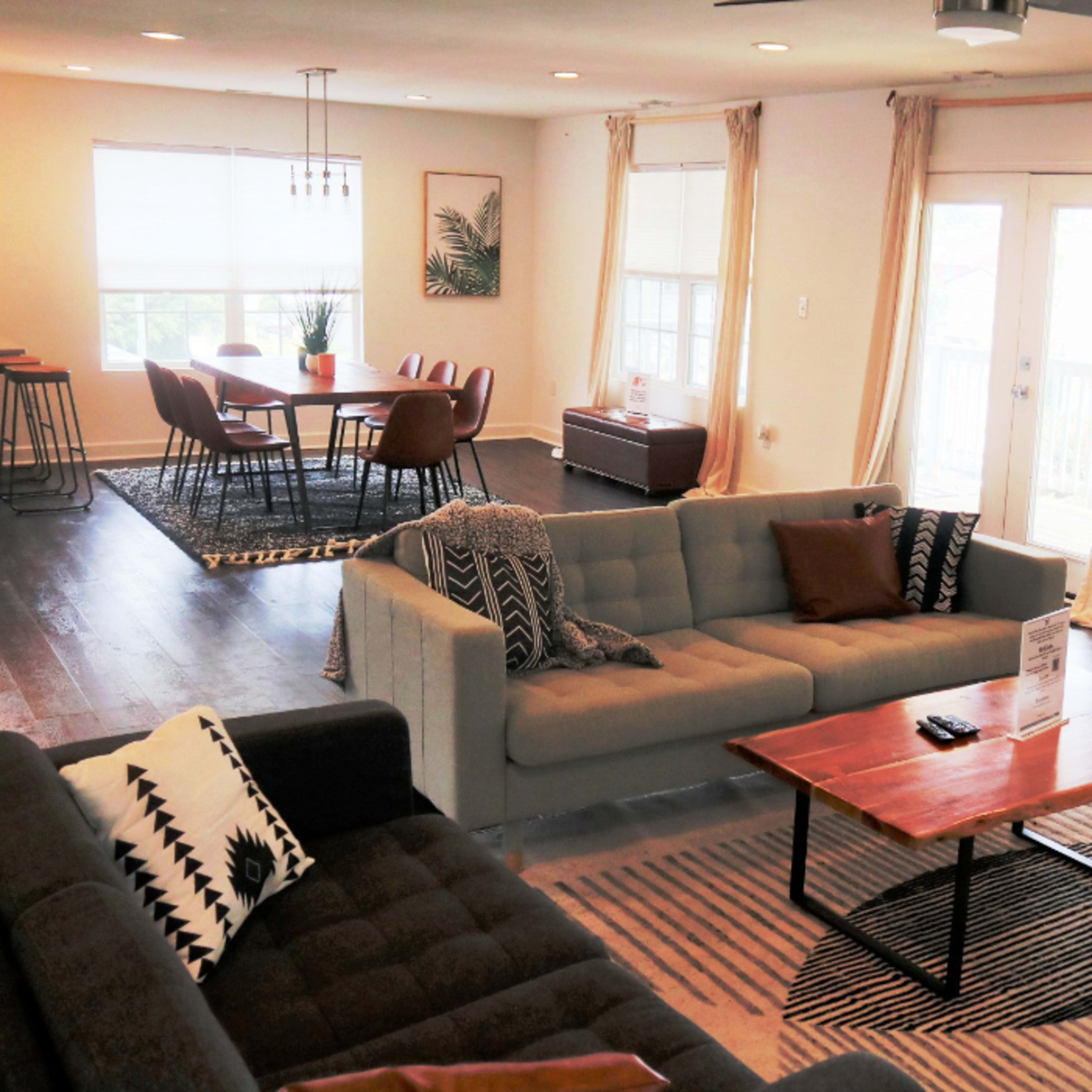 The living area features two sofas, a coffee table, and a dining table with chairs, all arranged in a bright, open space with large windows and wooden flooring.