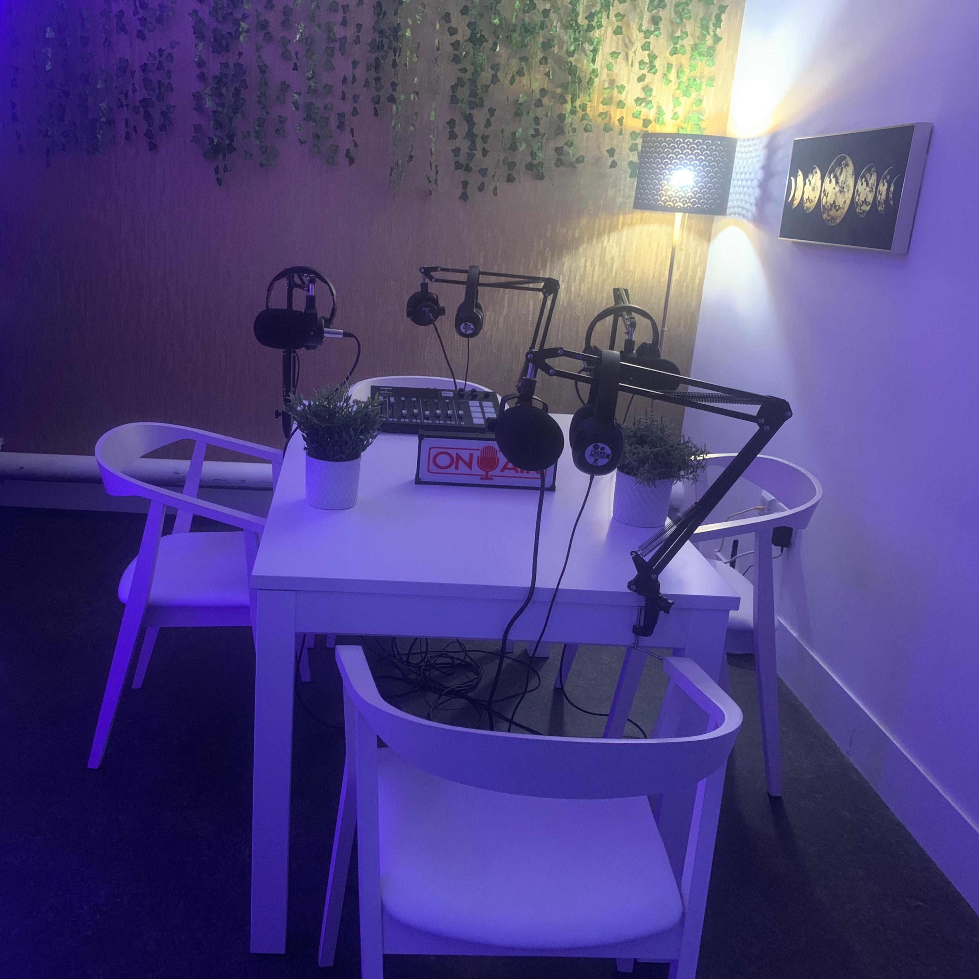 A small recording studio features a white table surrounded by chairs, microphones on stands, and decorative plants, with soft purple lighting.