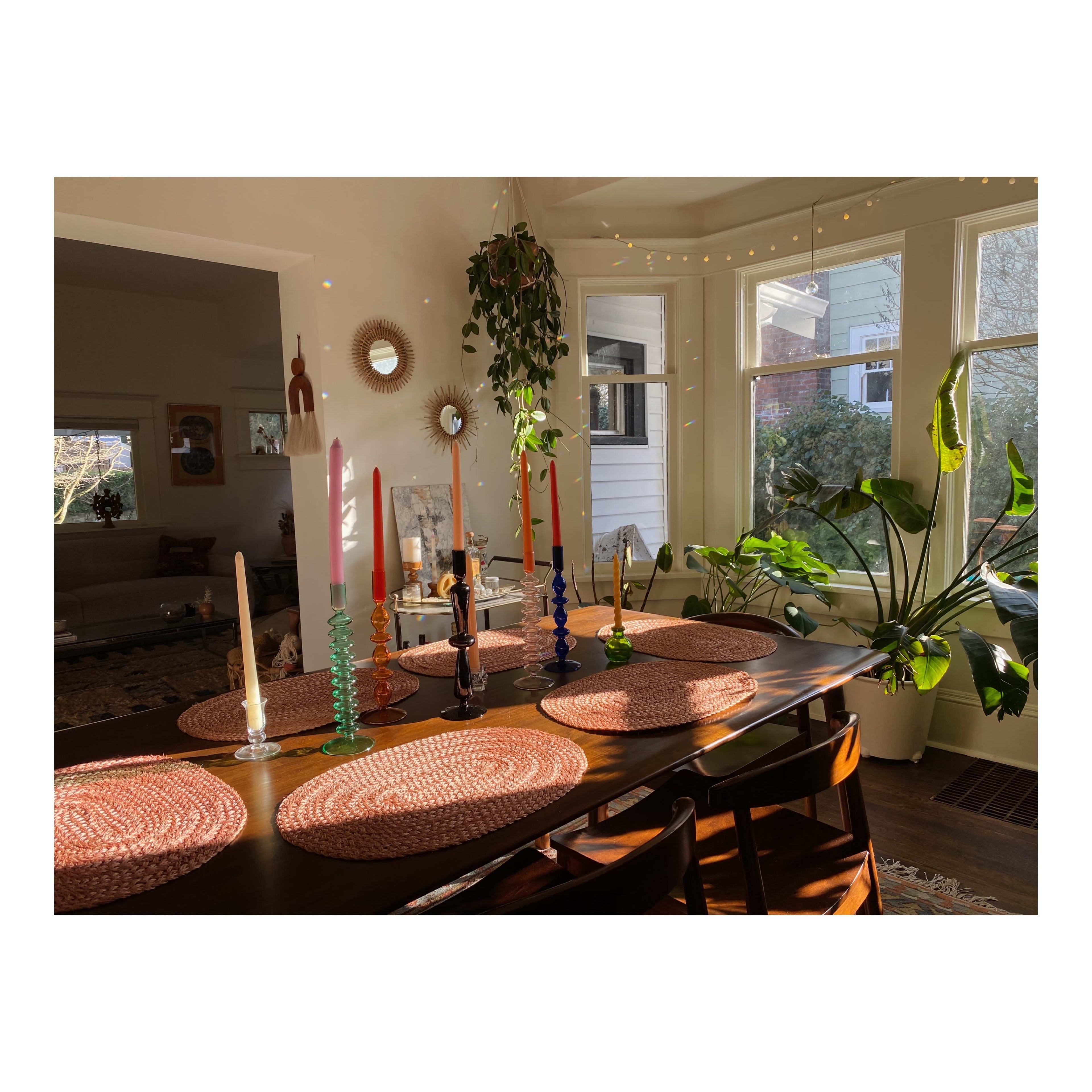 A dining table is set with colorful candles and woven placemats, illuminated by natural light from nearby windows.