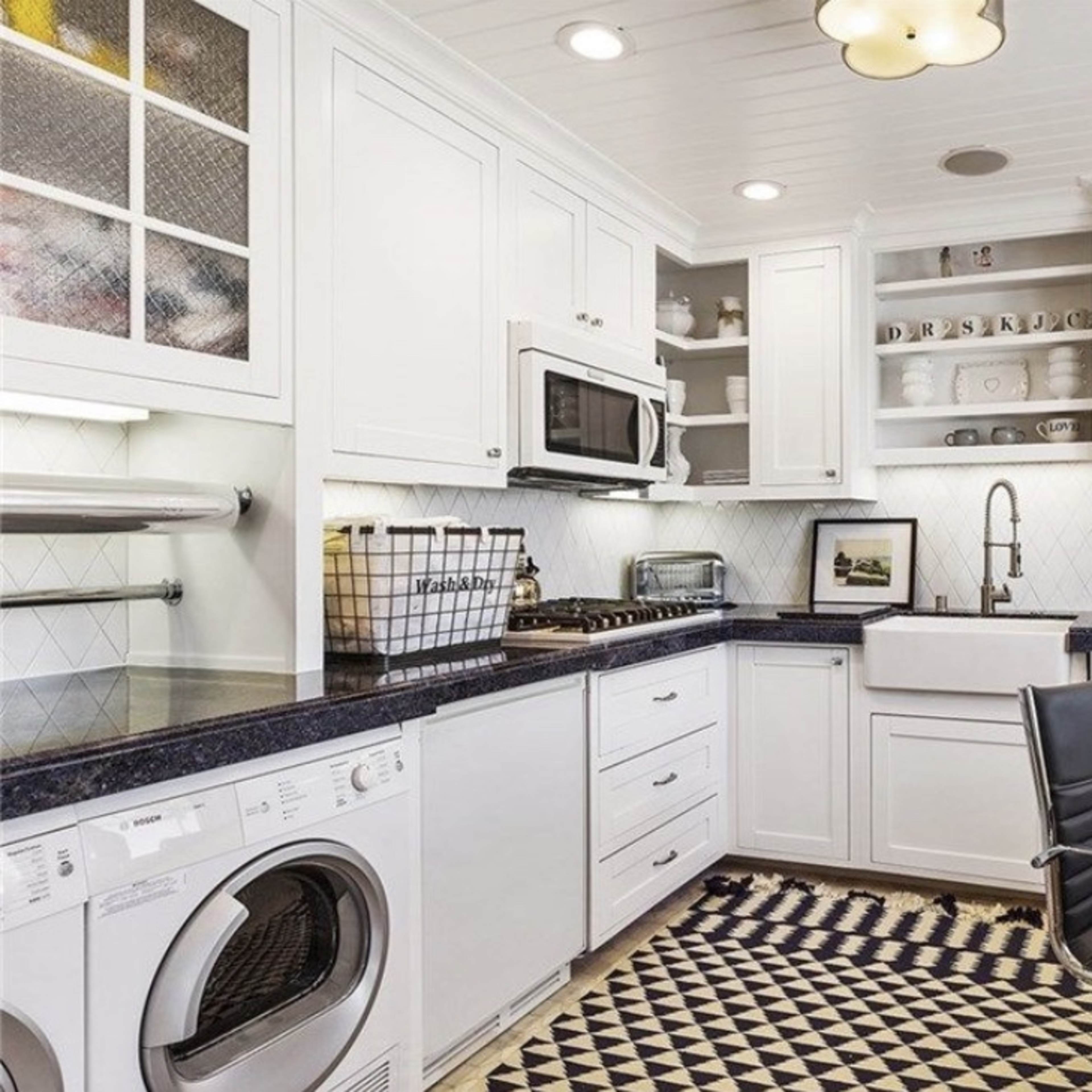 A modern laundry room featuring white cabinets, a black countertop, a farmhouse sink, and a washing machine, with a patterned rug and a black chair.