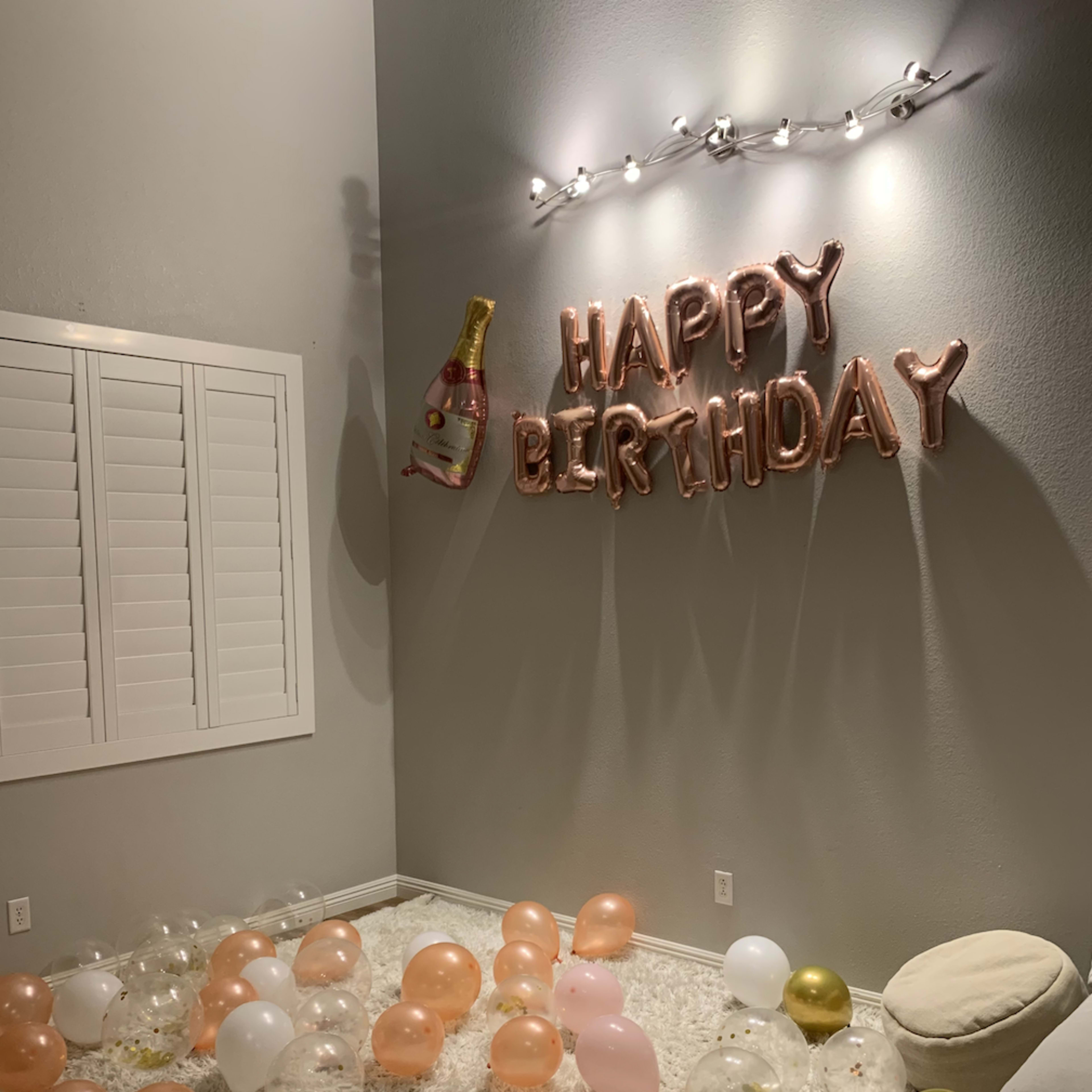 A celebration setting features a "HAPPY BIRTHDAY" balloon banner, a champagne bottle decoration, and a variety of balloons scattered on a white rug.