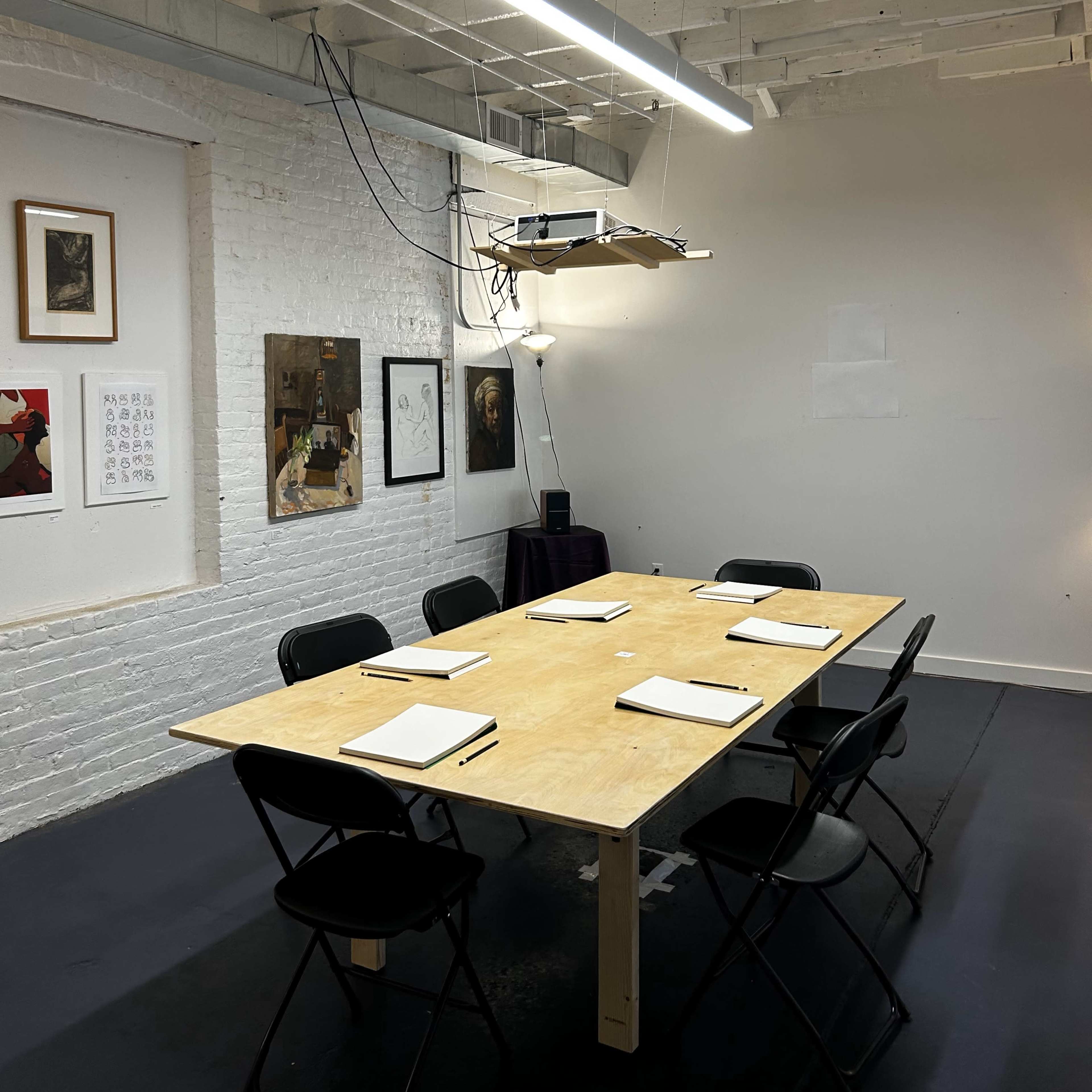 A spacious, minimalist meeting room features a wooden table surrounded by black chairs, with overhead lighting and artwork on the white brick walls.