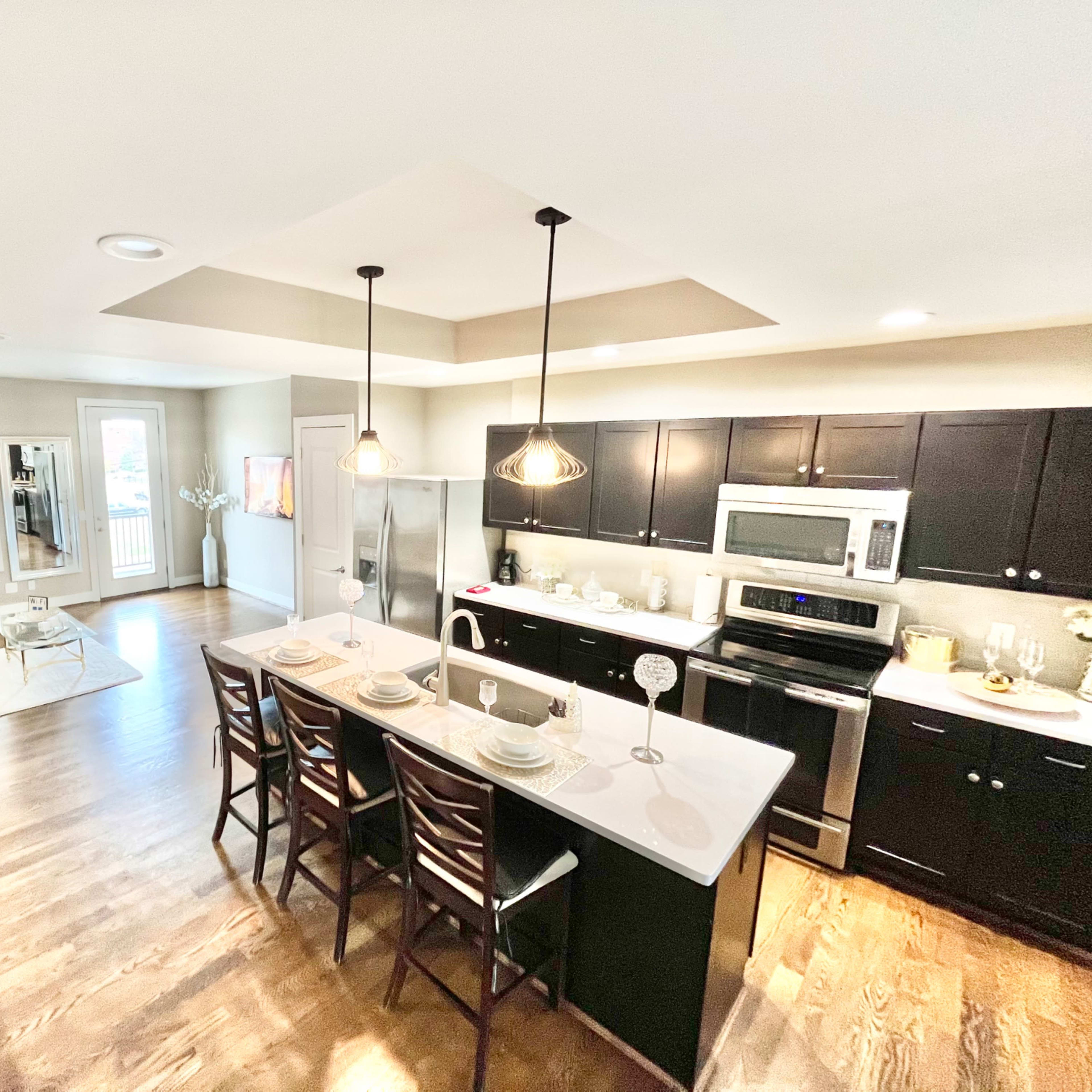 A modern kitchen with dark cabinetry, stainless steel appliances, and a central island featuring four bar stools, overlooking a living area with natural light.