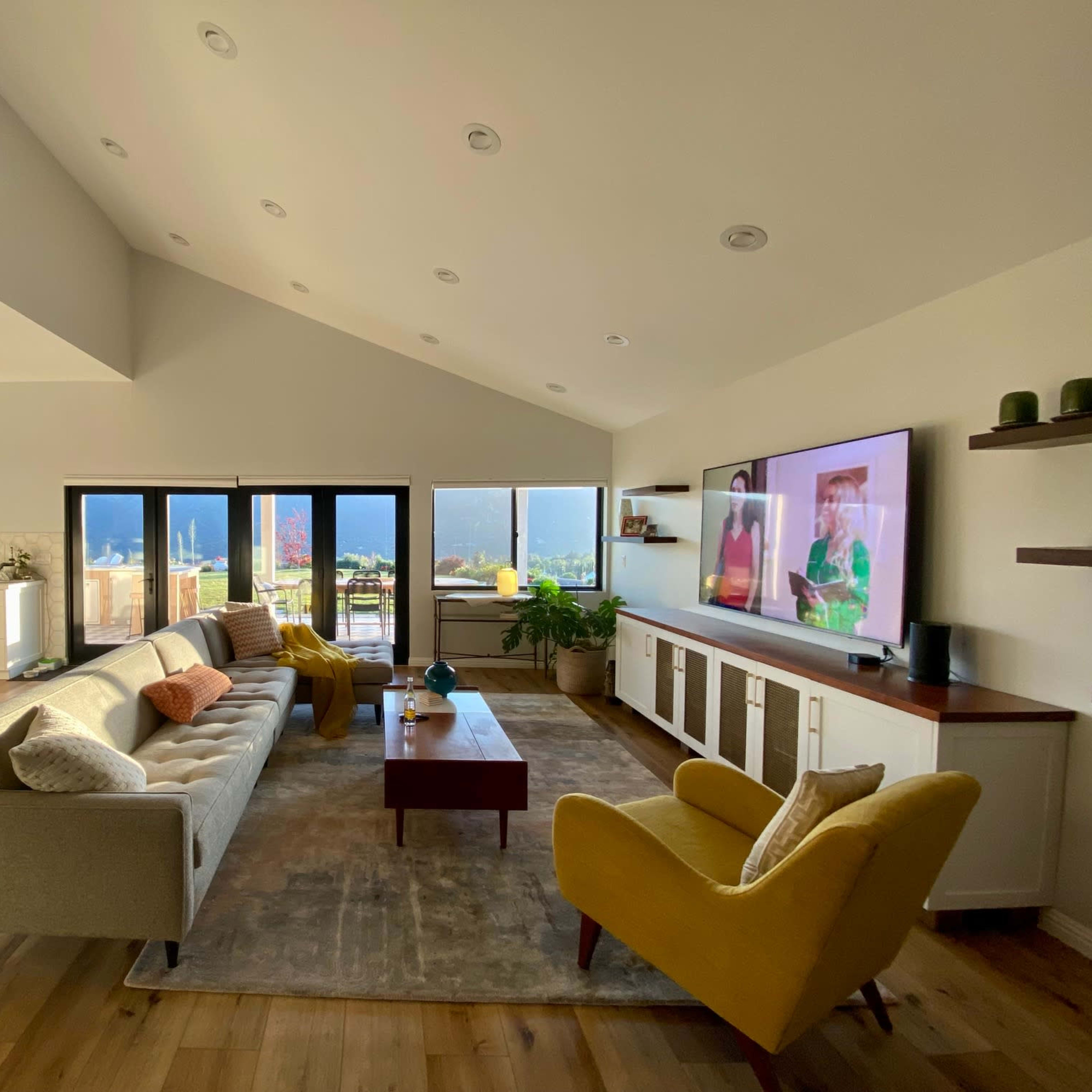 A modern living room features a sectional sofa, an accent chair, a large television, and expansive windows that provide a view of the outdoors.