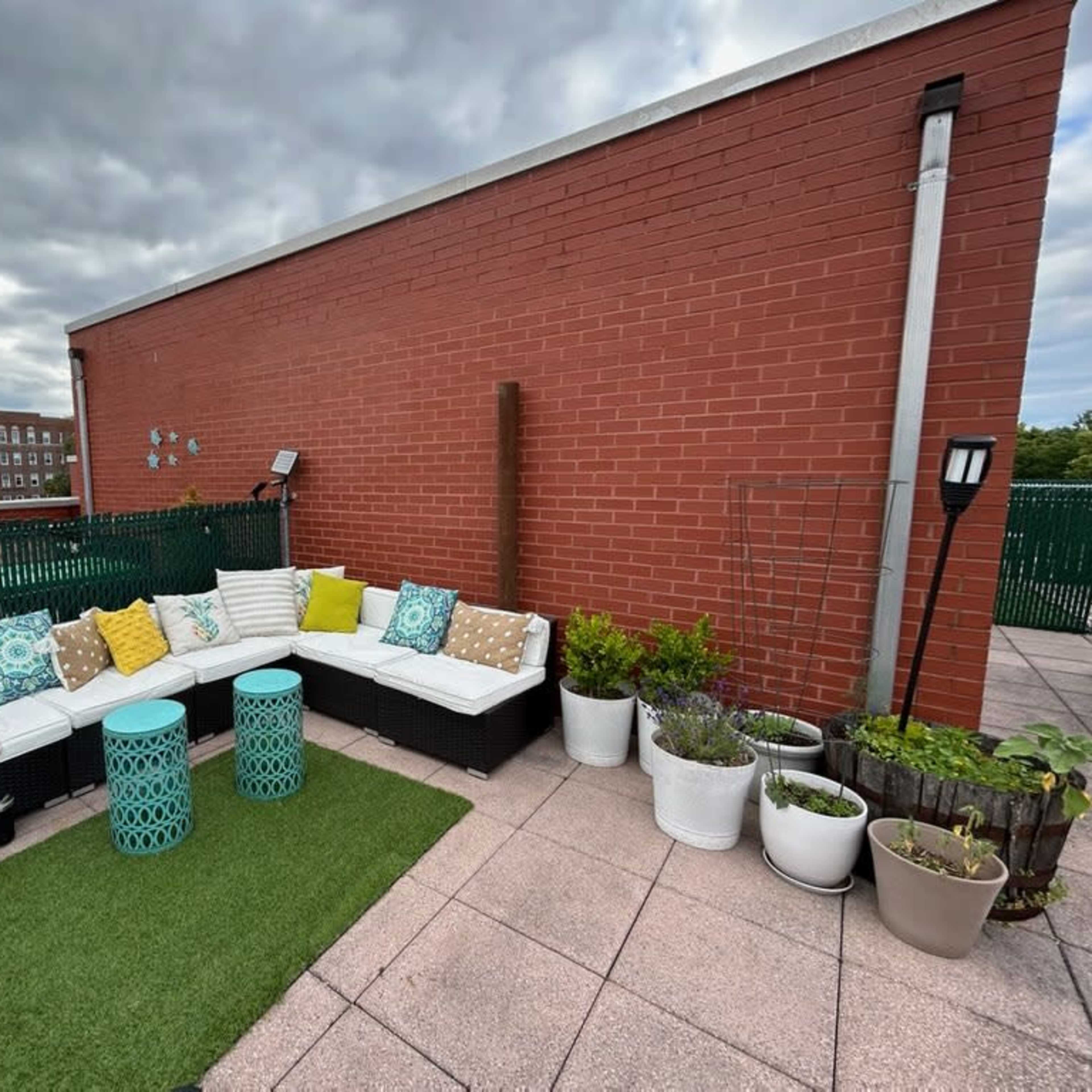 A rooftop patio features a sectional couch with colorful cushions, two turquoise tables, and various potted plants set on a green rug.