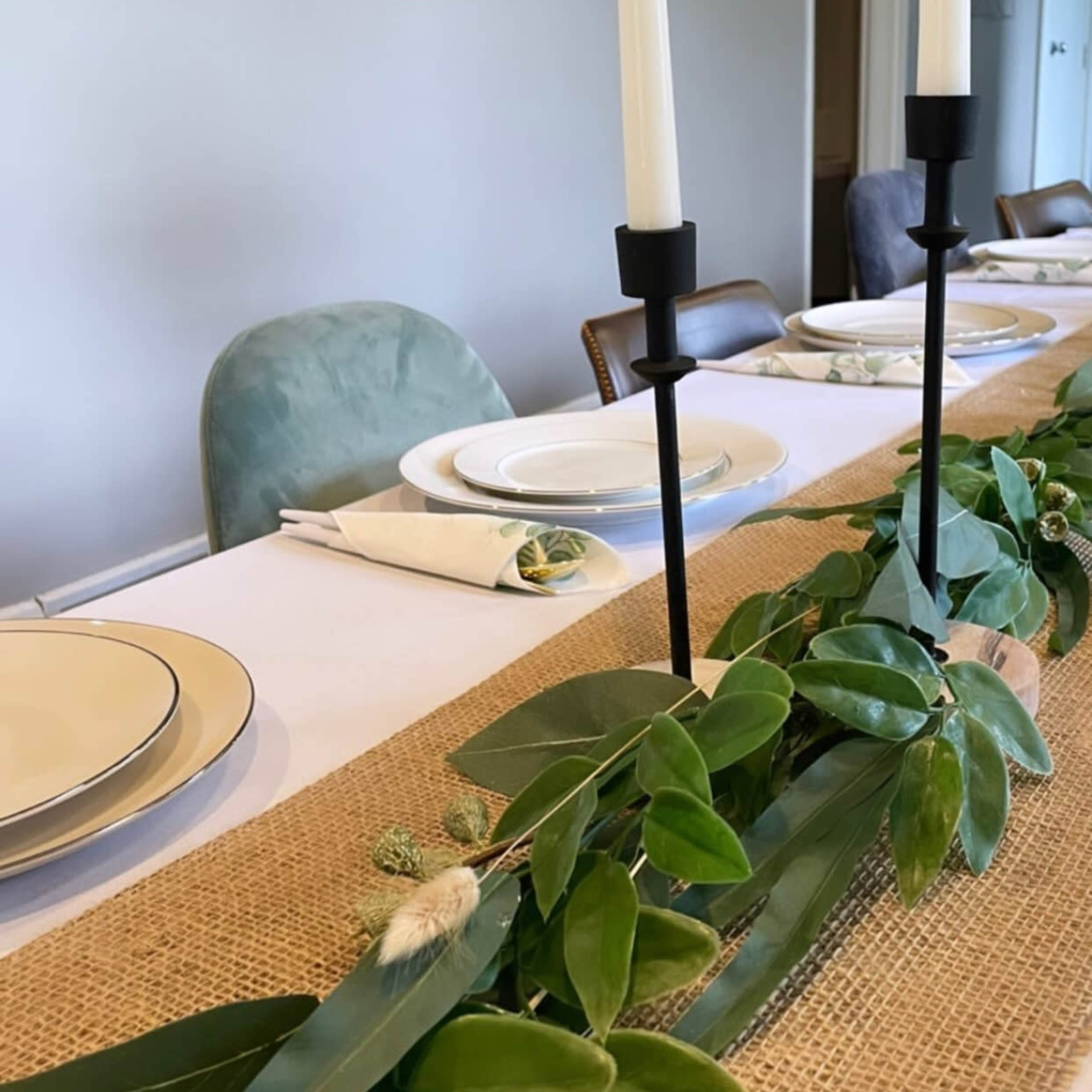 A long dining table is set with white plates and green plants, featuring tall candlesticks illuminating the centerpiece of greenery.