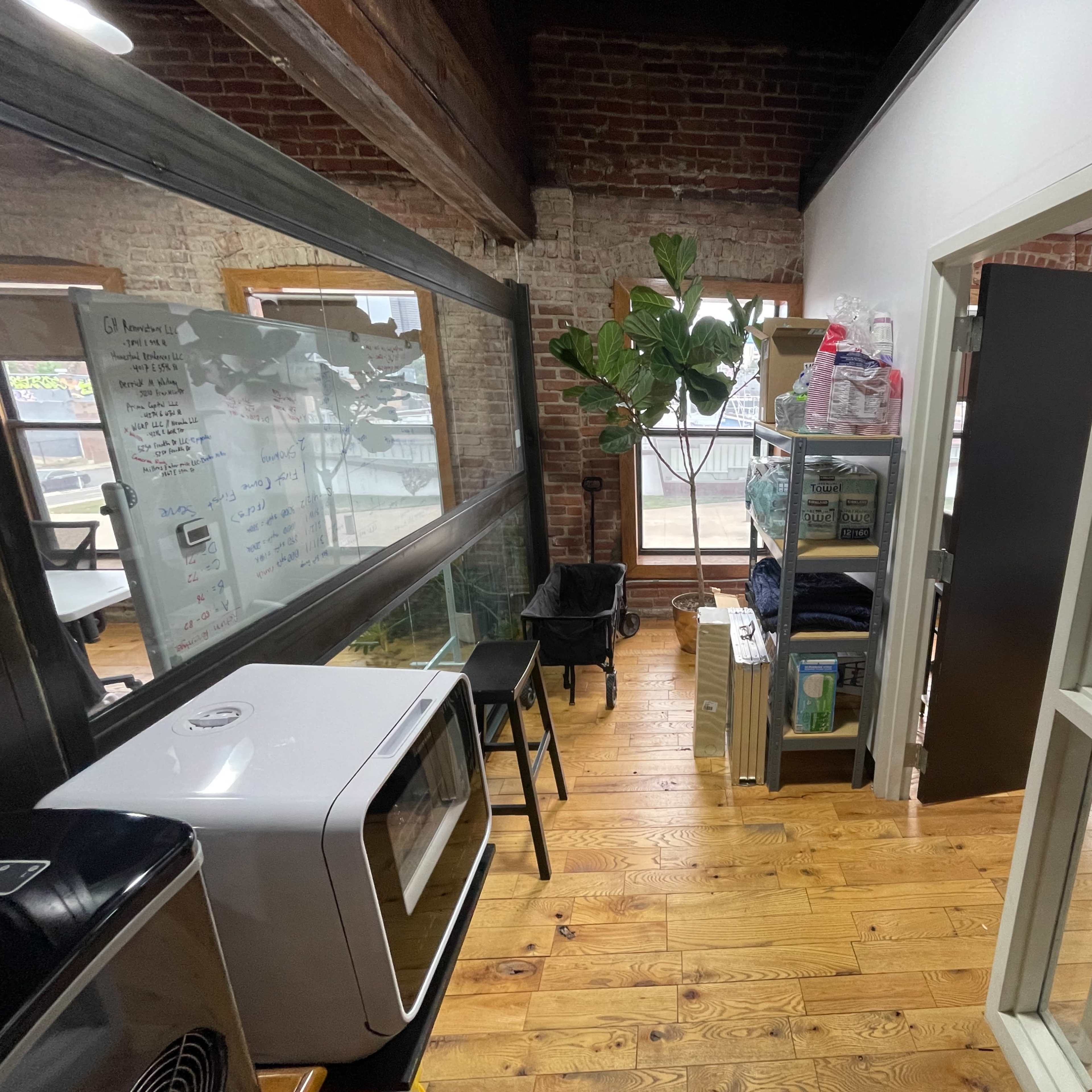 A small office space features a kitchenette area with a refrigerator and seating, alongside a shelf filled with supplies and a large window providing natural light.
