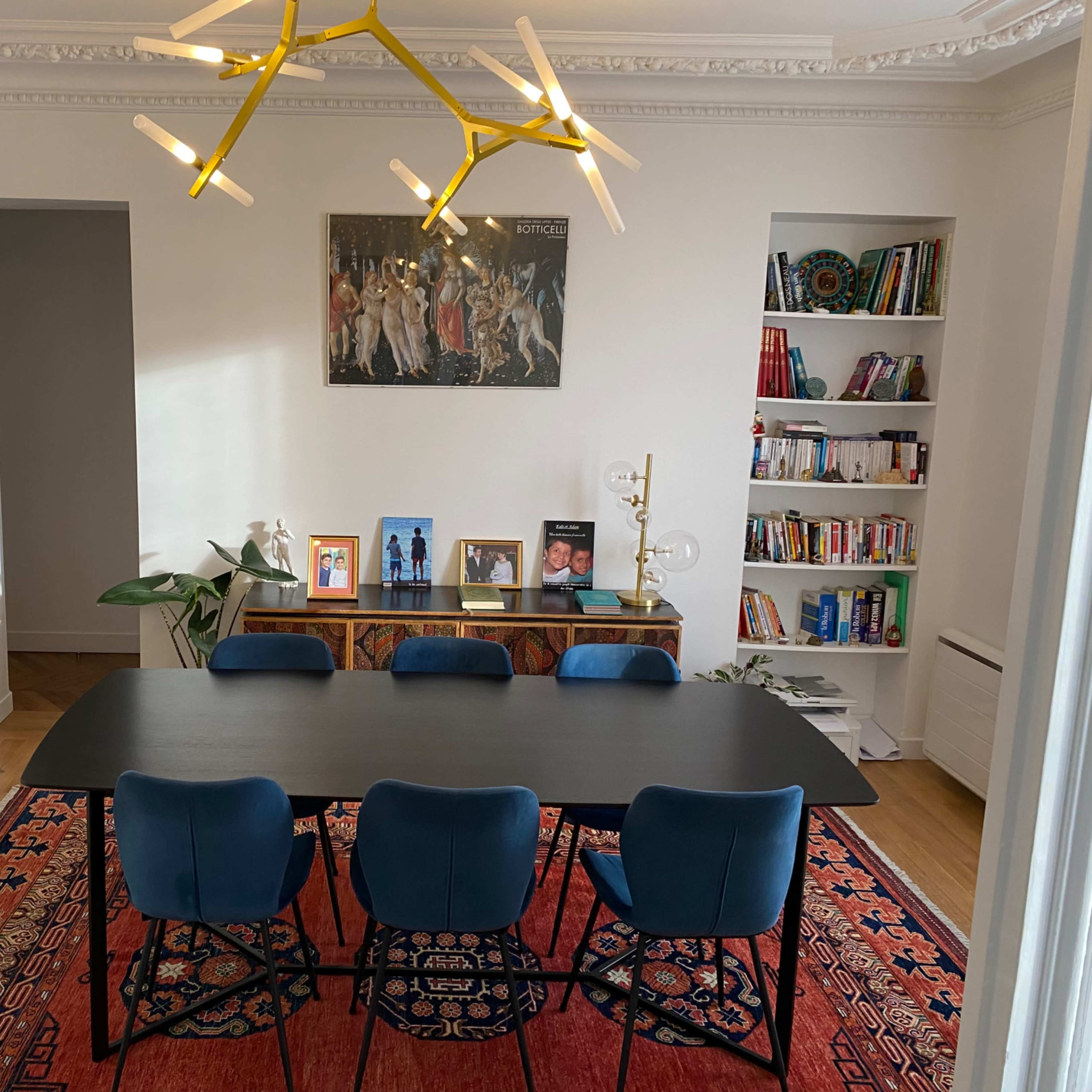 A modern dining area with a large black table surrounded by blue chairs, a colorful rug, and shelves filled with books and decorative items.