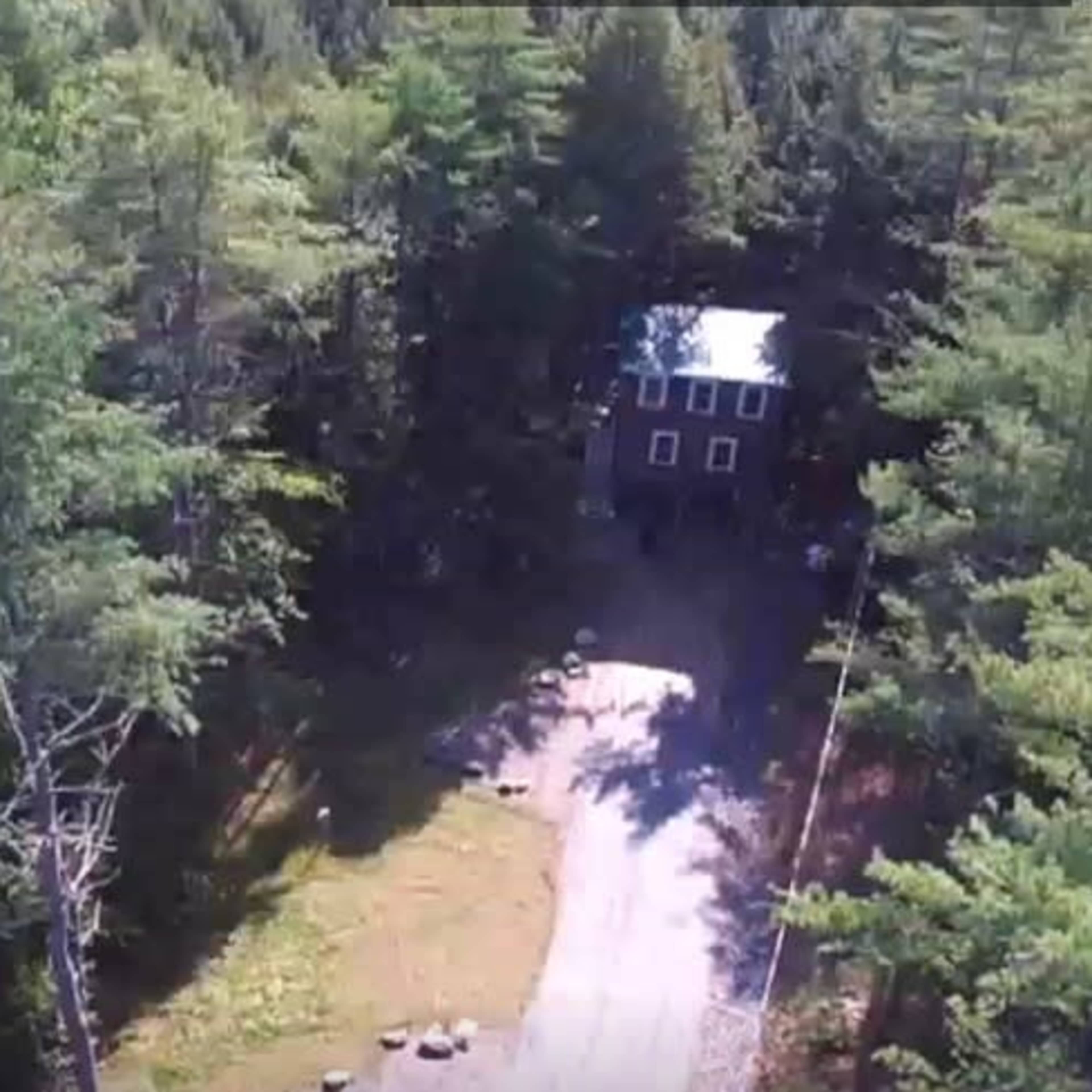 A two-story house is situated at the end of a gravel driveway, surrounded by dense trees.