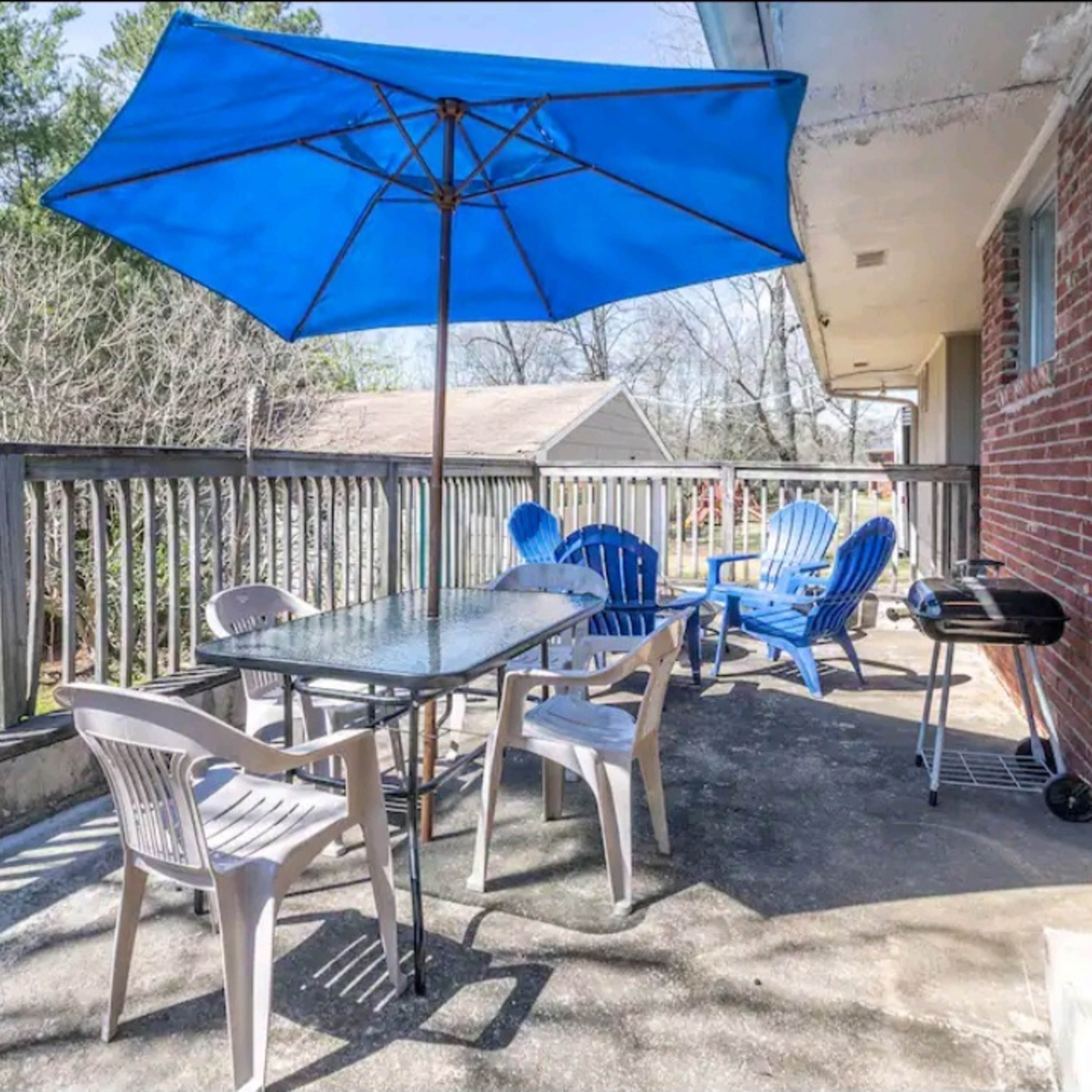 A patio features a glass-top table surrounded by white chairs, an umbrella, and several blue lounge chairs, with a barbecue grill nearby.