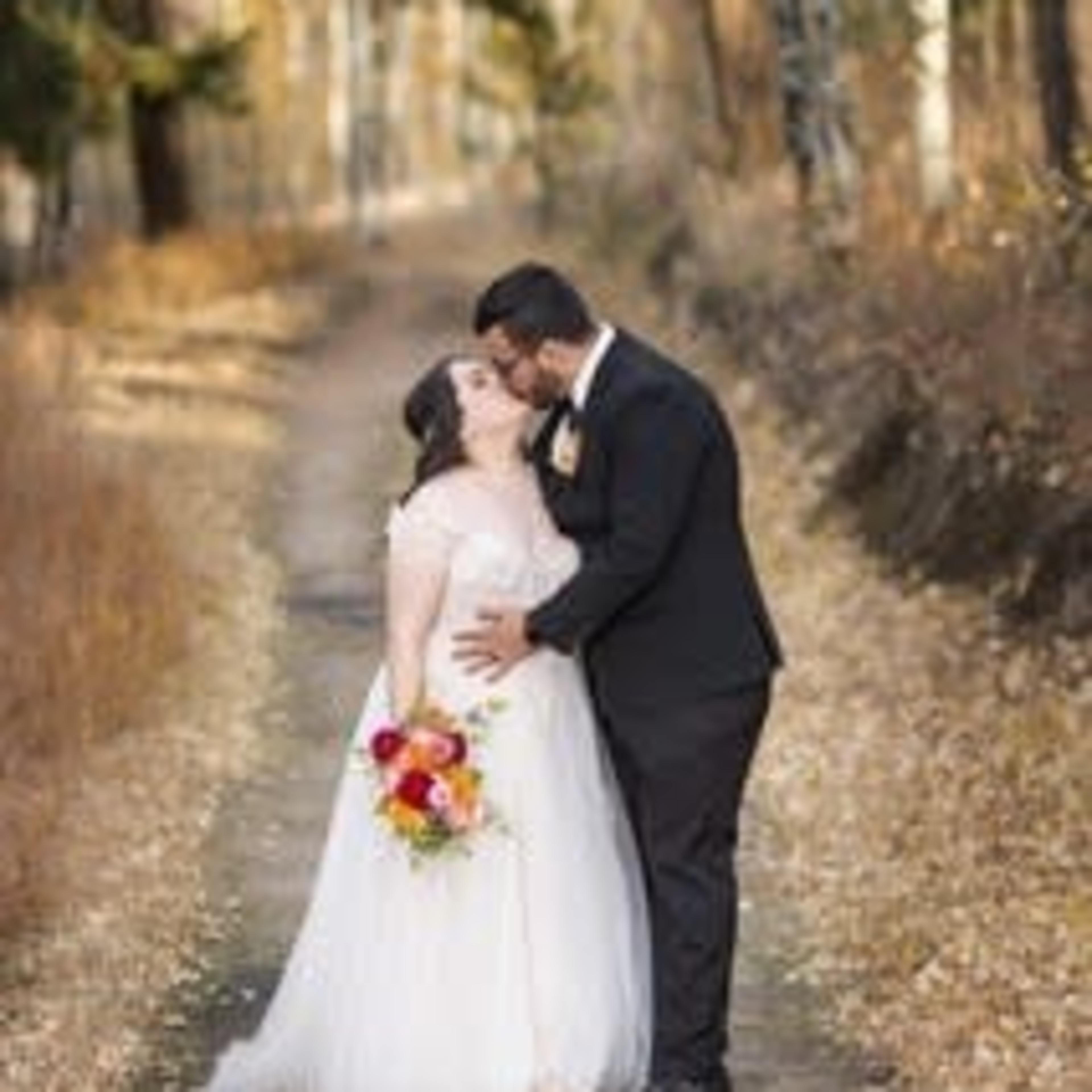 A couple stands on a leaf-covered path, sharing a kiss while the bride holds a bouquet of flowers.