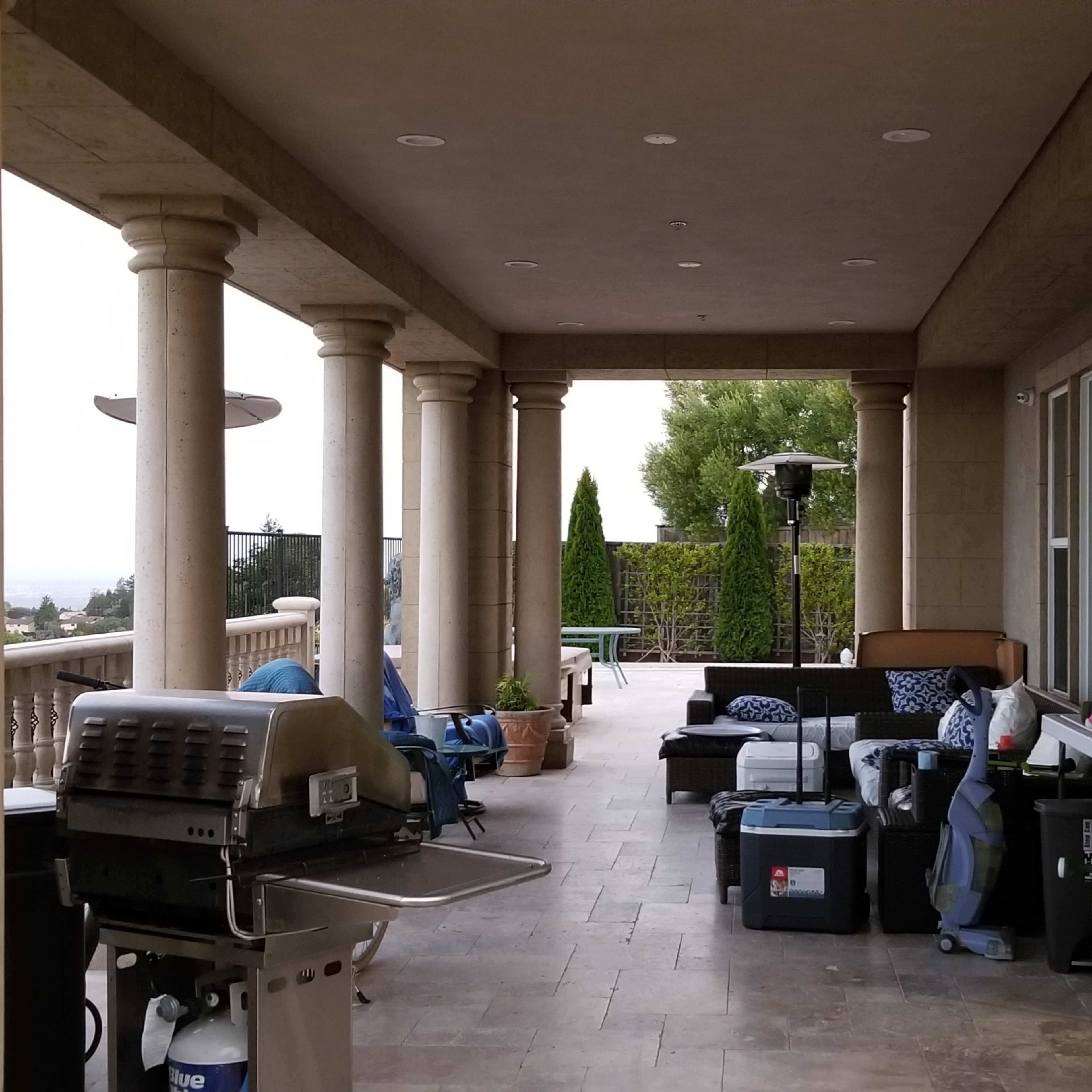 A covered patio with columns features a barbecue grill and various boxes and furniture positioned along the edges.