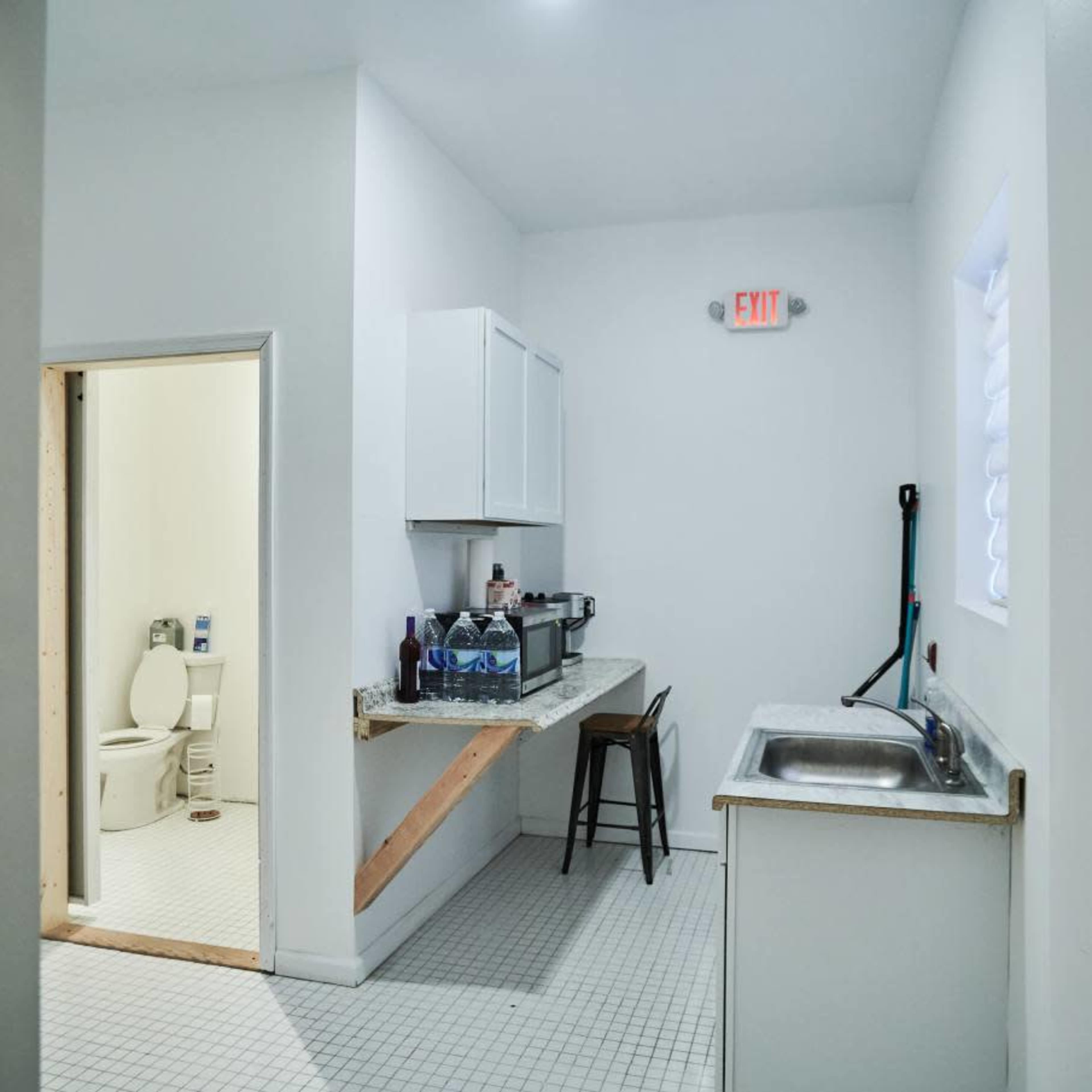 A small, minimalist laundry room with a sink, countertop, and an adjoining bathroom visible through an open doorway.