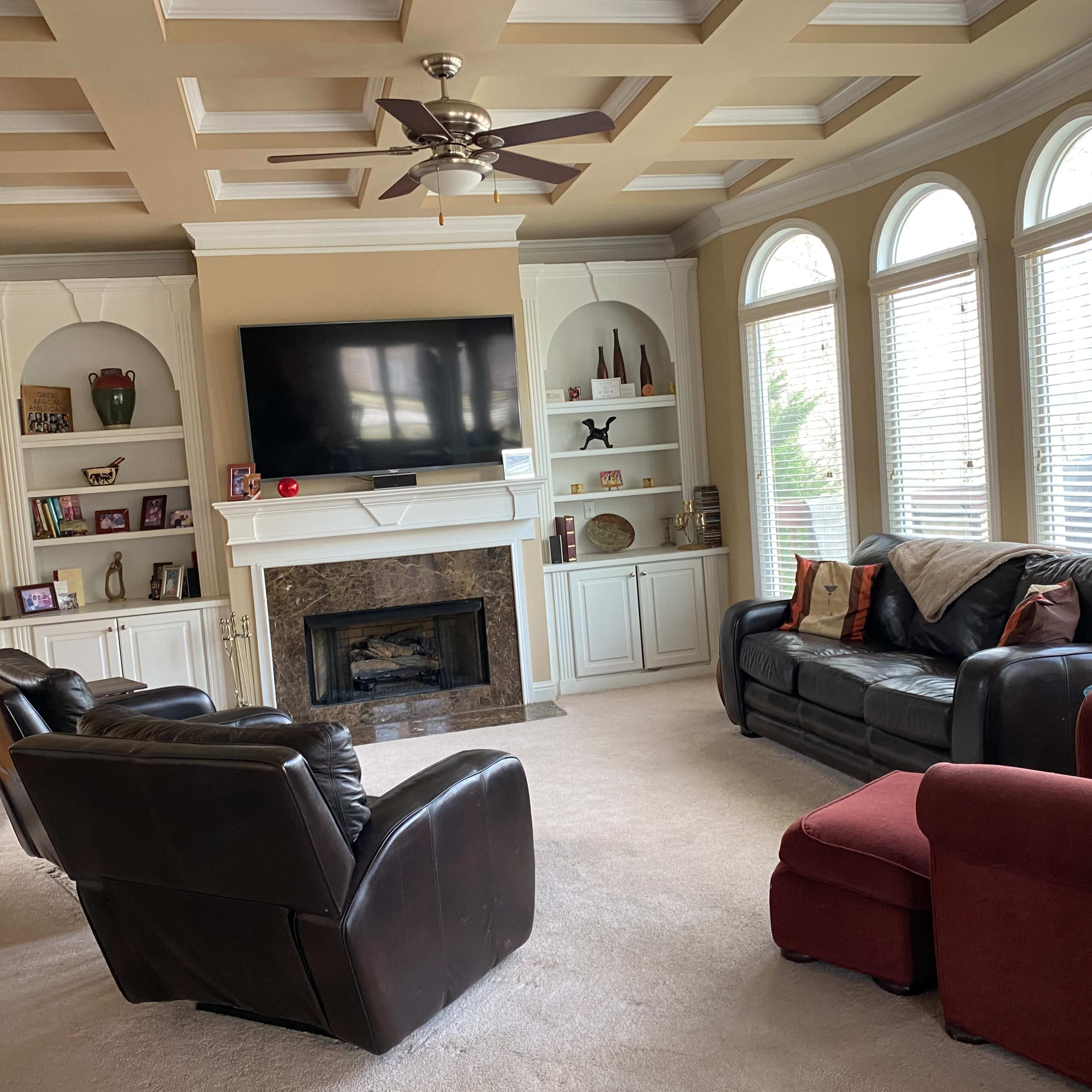 A living room features a coffered ceiling, a television mounted above a fireplace, and three seating arrangements, including a red chair, in front of large windows.
