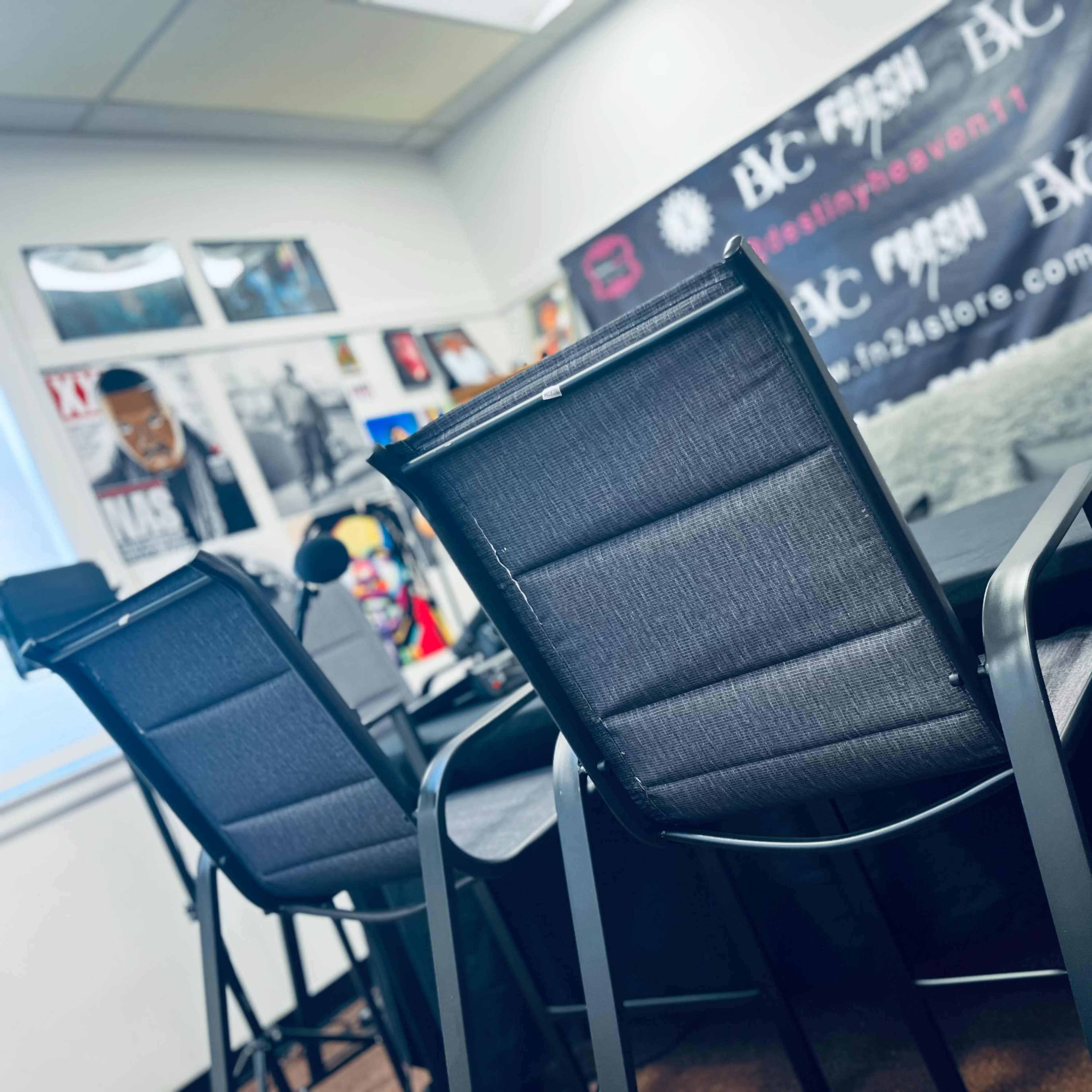 A room setup for a podcast or interview, featuring a table with two chairs, microphones, and a backdrop with various posters and branding.