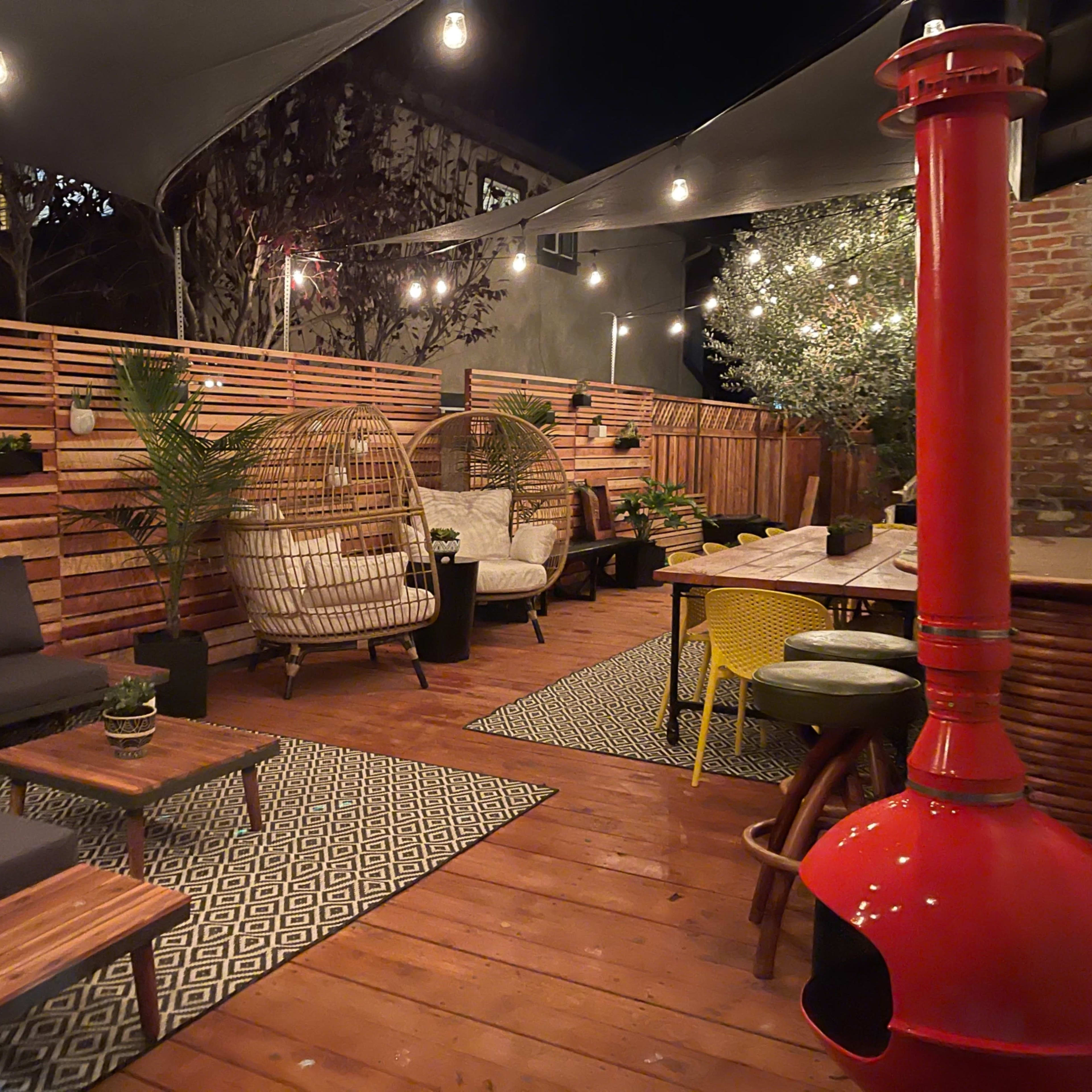 A cozy outdoor seating area with wooden decking, plants, and string lights, featuring comfortable furniture arranged around a central table and a red outdoor stove.