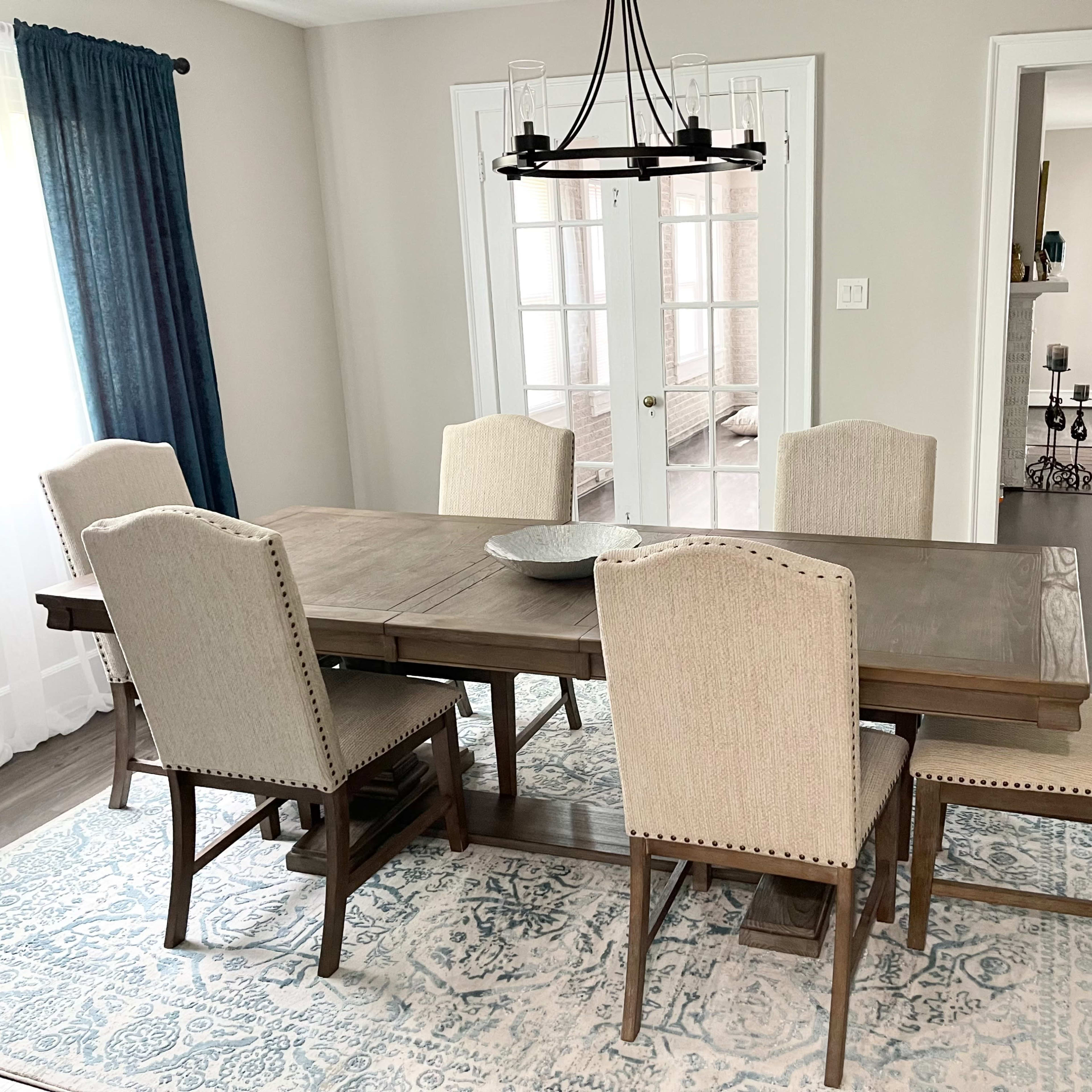 A wooden dining table with six upholstered chairs is set on a patterned rug in a well-lit dining room, featuring large windows and a chandelier above the table.